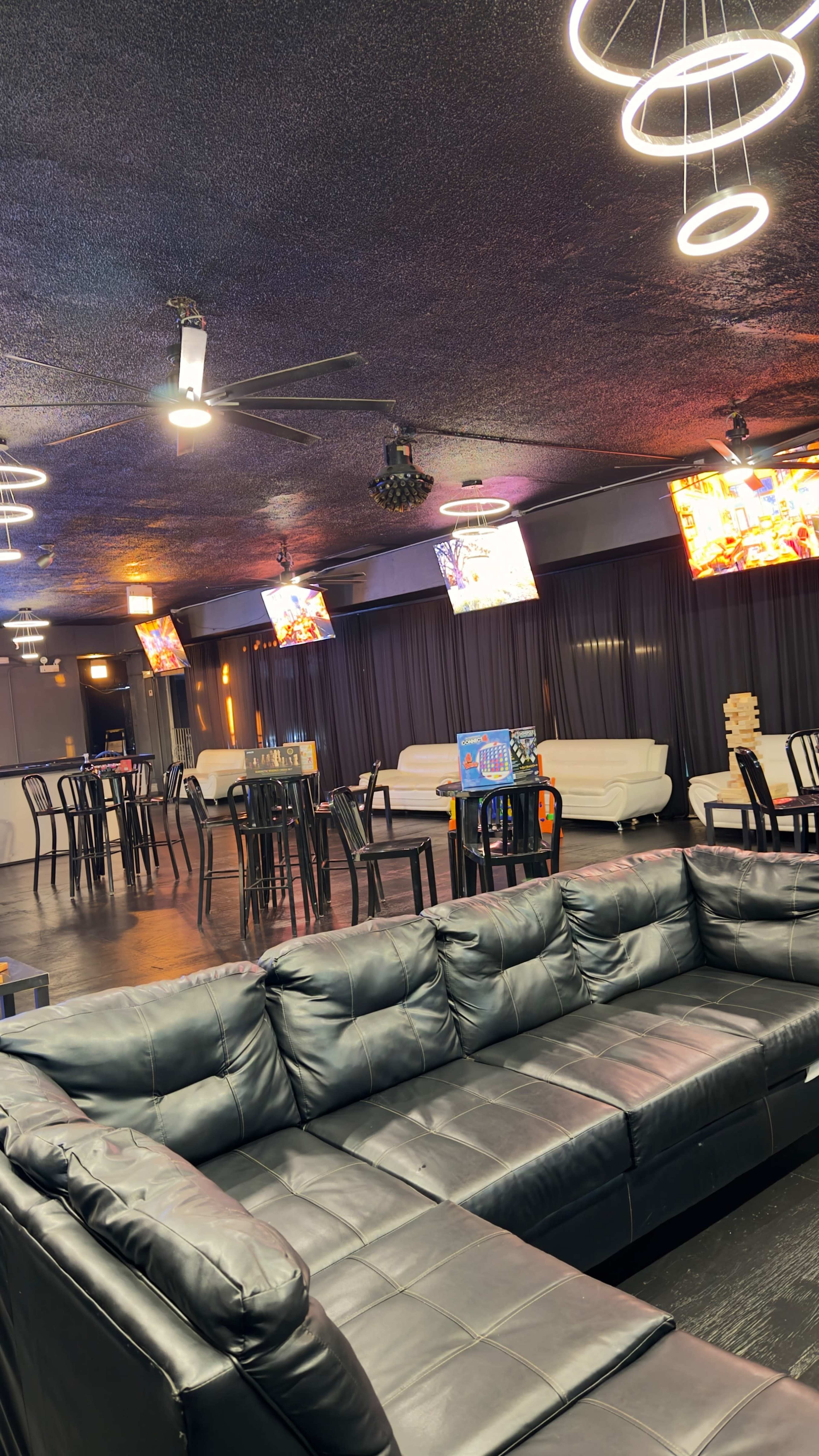 The image shows a modern lounge area with black couches, several dining tables, and multiple screens displaying colorful visuals.