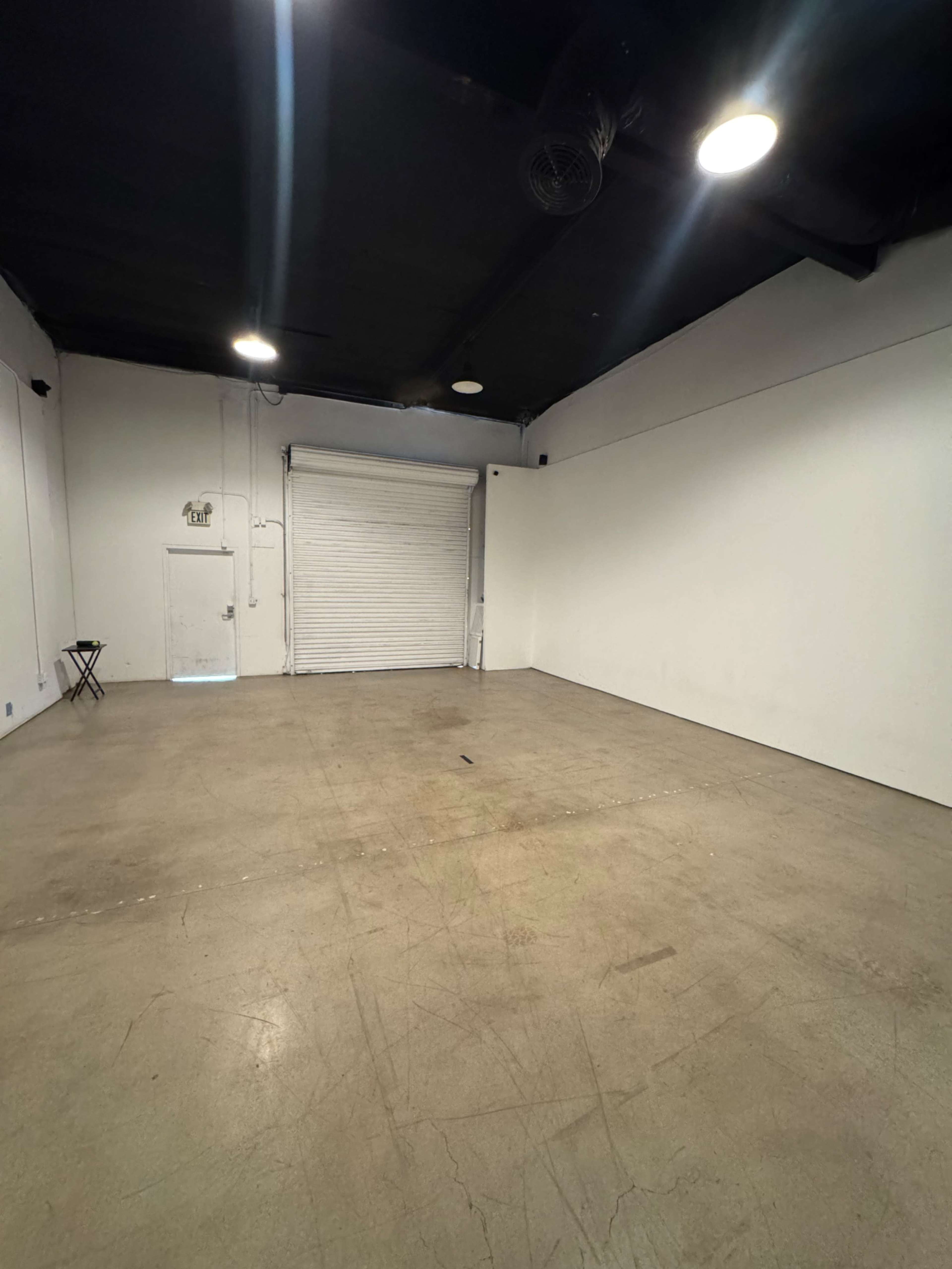 The image shows an empty room with a concrete floor, a white wall, and a closed roll-up door at one end.
