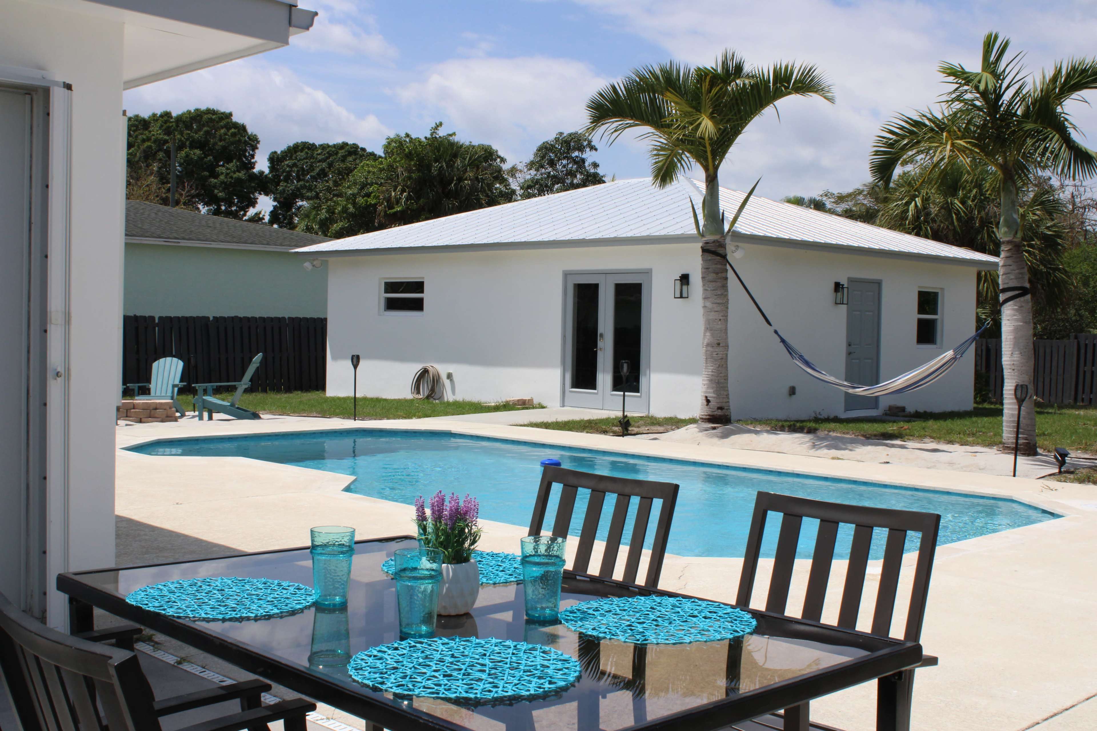 A backyard with a swimming pool, outdoor dining table, and a hammock hanging between two palm trees beside a white shed.