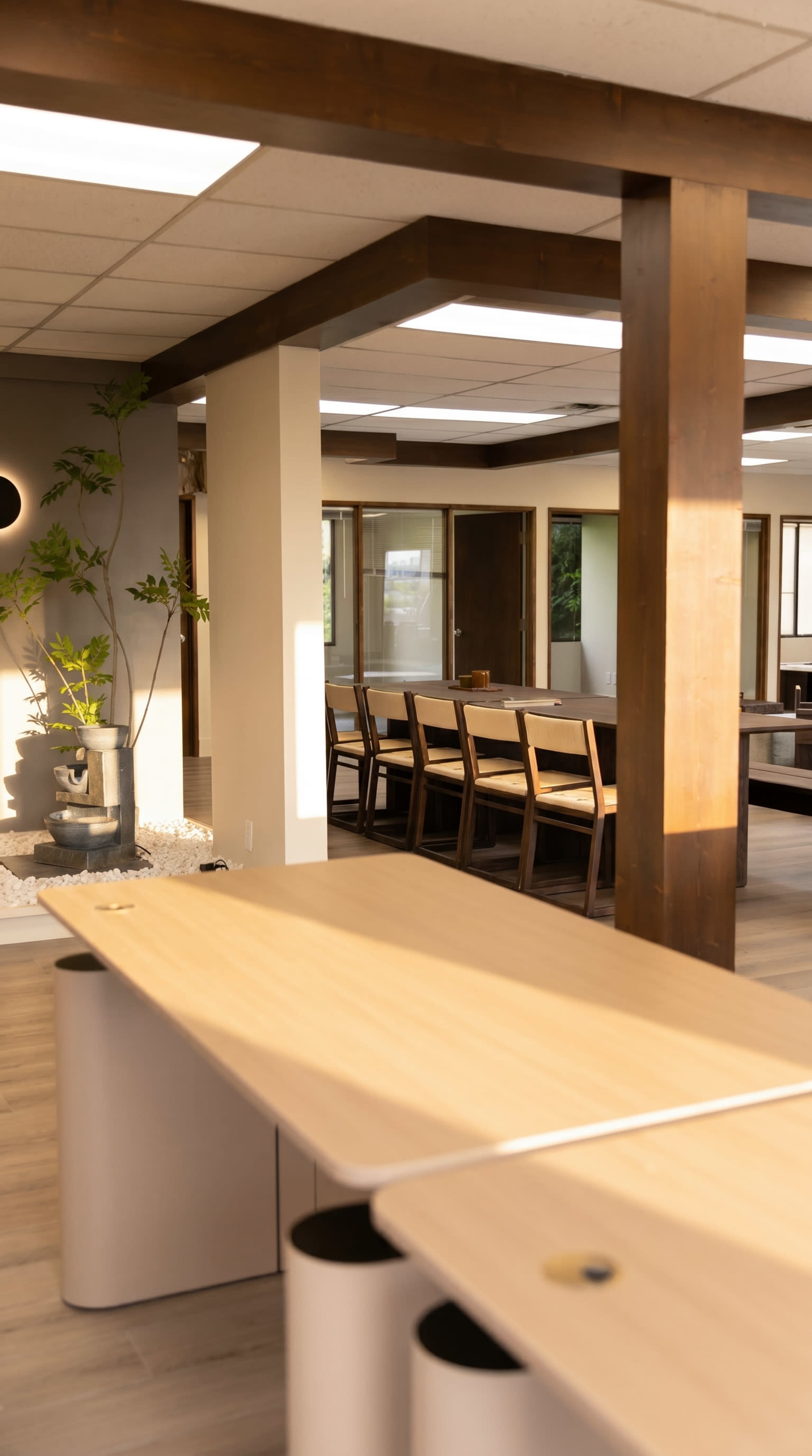 The image shows a modern office interior with wooden accents, a large communal table, and a seating area with plants and a water feature.