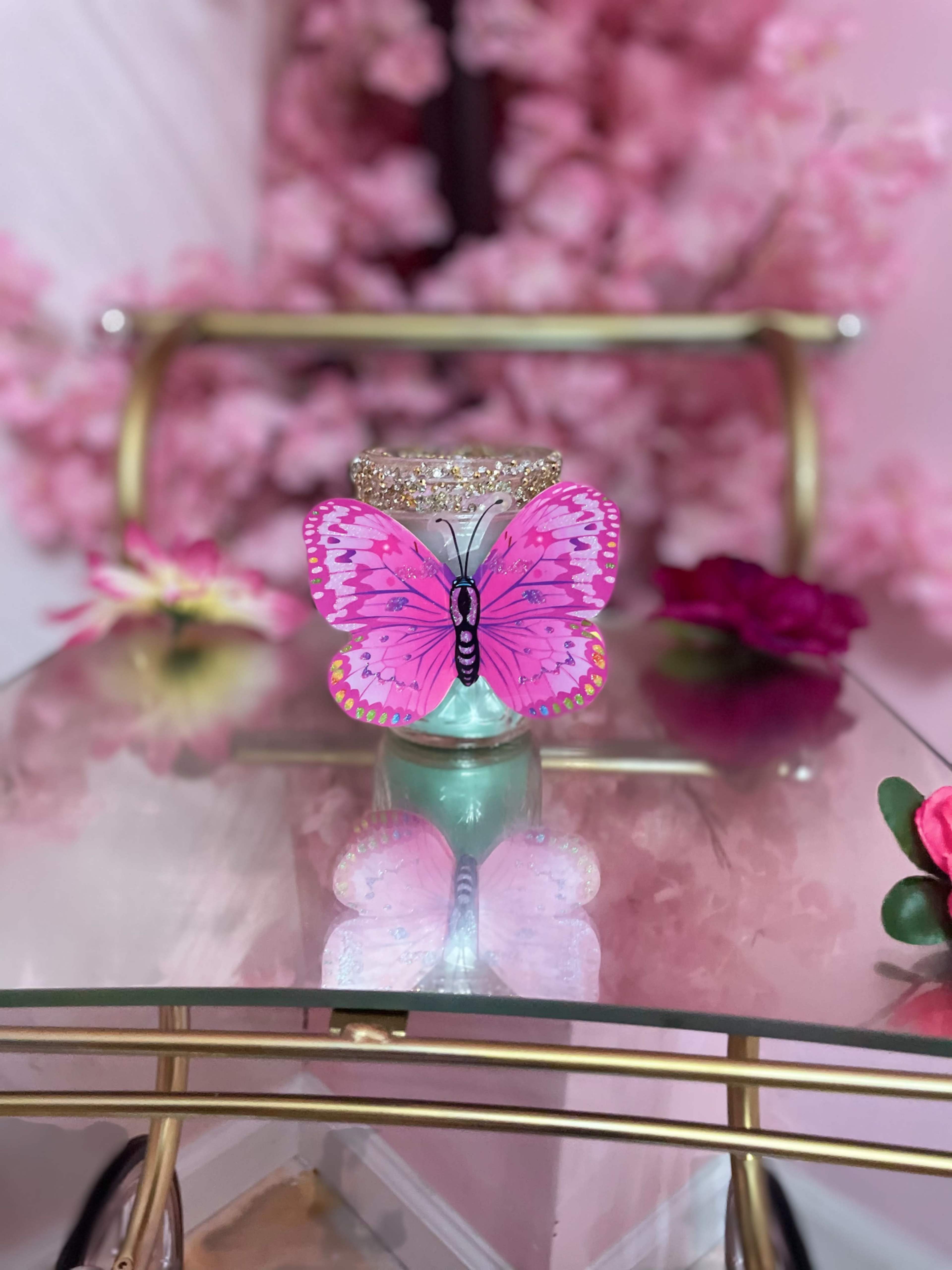 A glass container adorned with a pink butterfly sits on a glass table surrounded by decorative flowers and cherry blossom branches.
