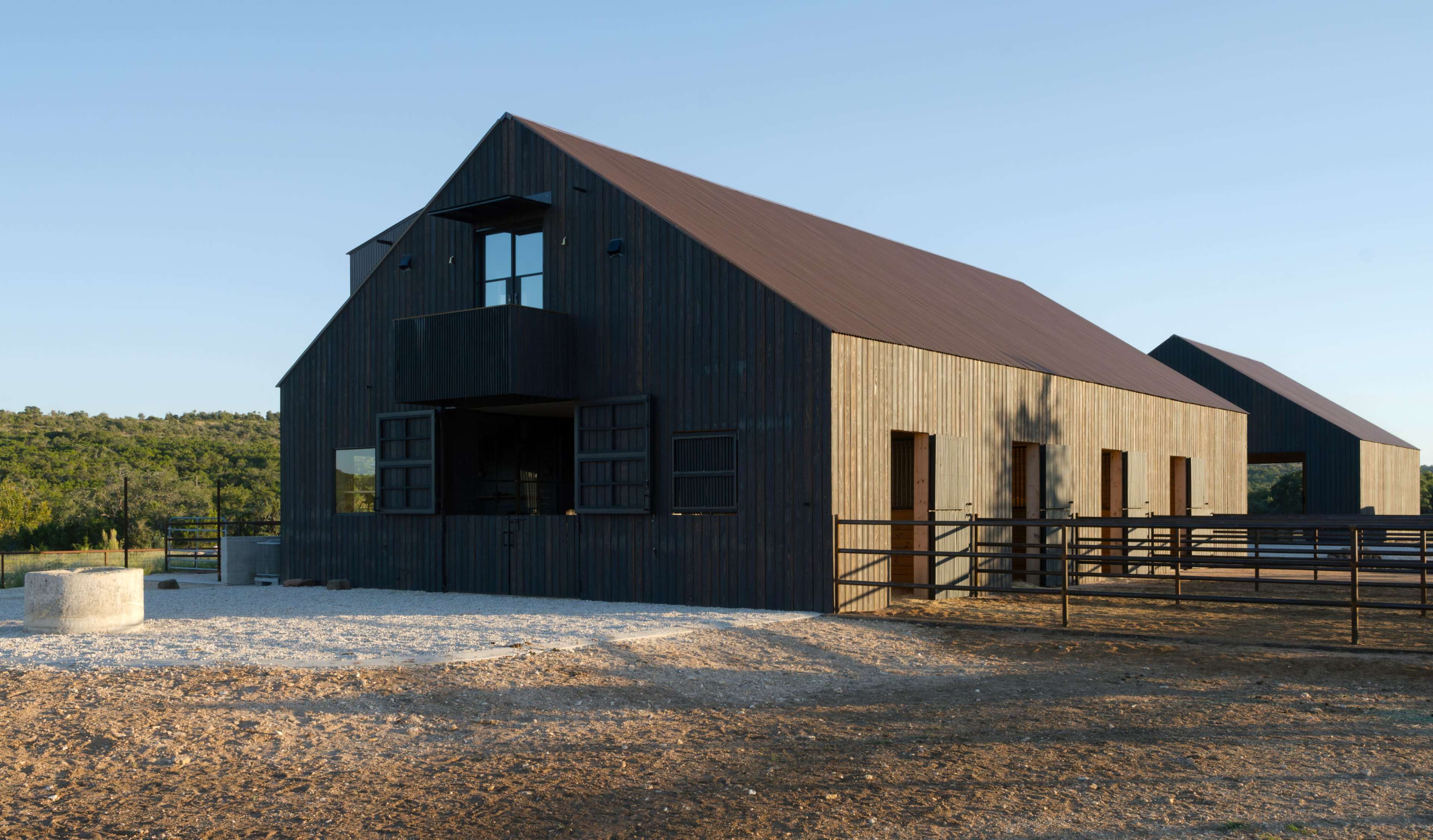 A large, modern barn with a dark wooden exterior and a metal roof stands in an open area surrounded by greenery.