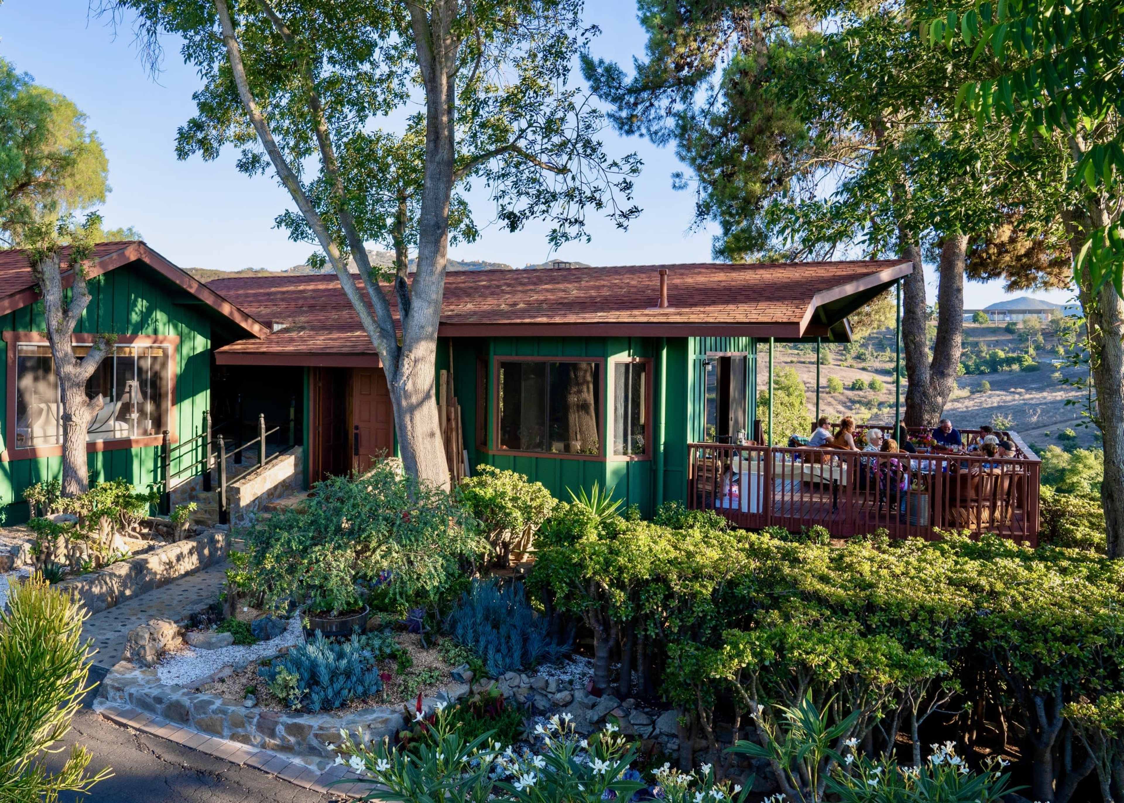 A green house with a red roof features a deck where people are seated, surrounded by trees and well-maintained landscaping.