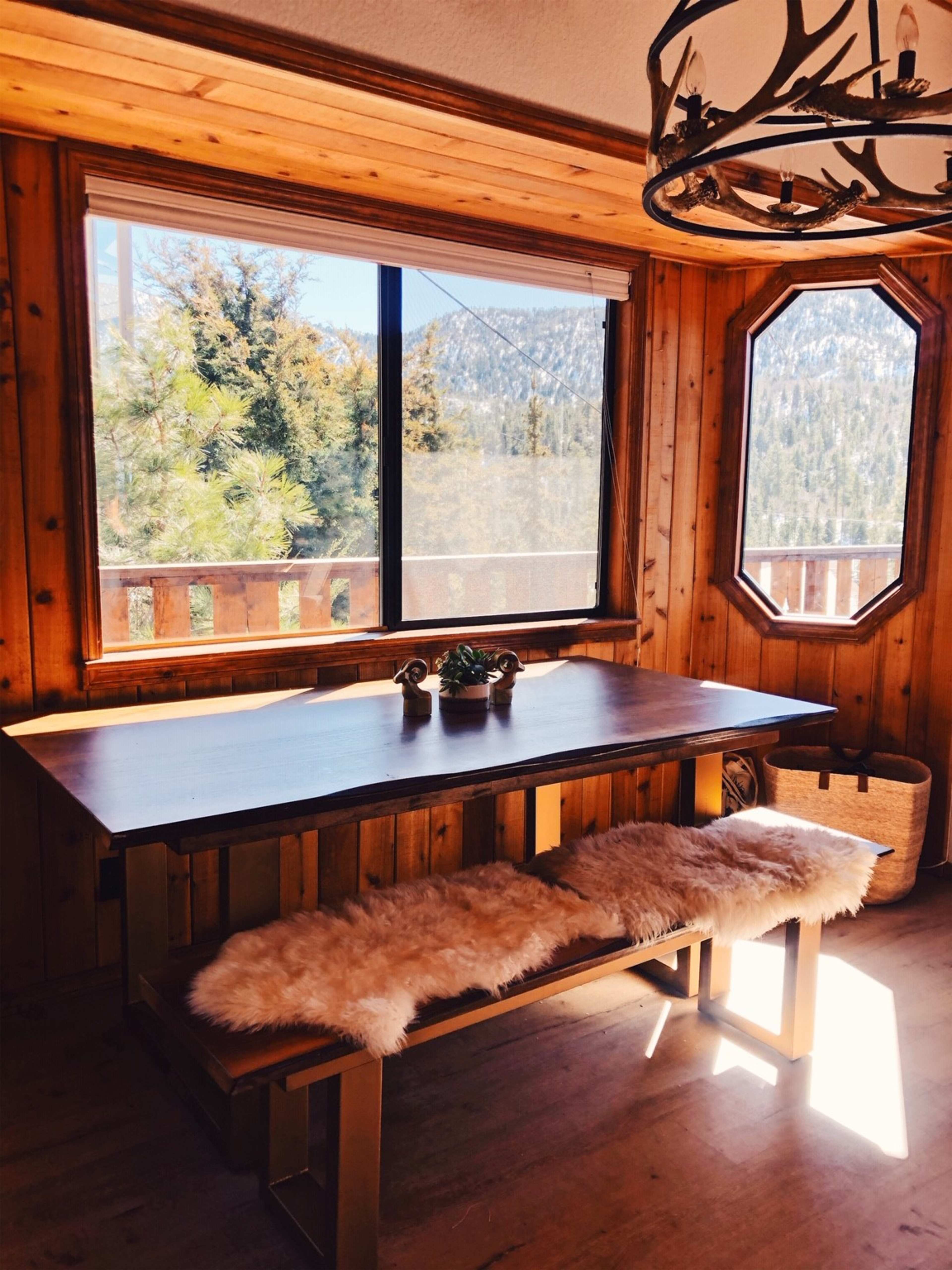 A wooden dining table with a bench covered in fur sits in a sunlit room with large windows displaying a mountain view.