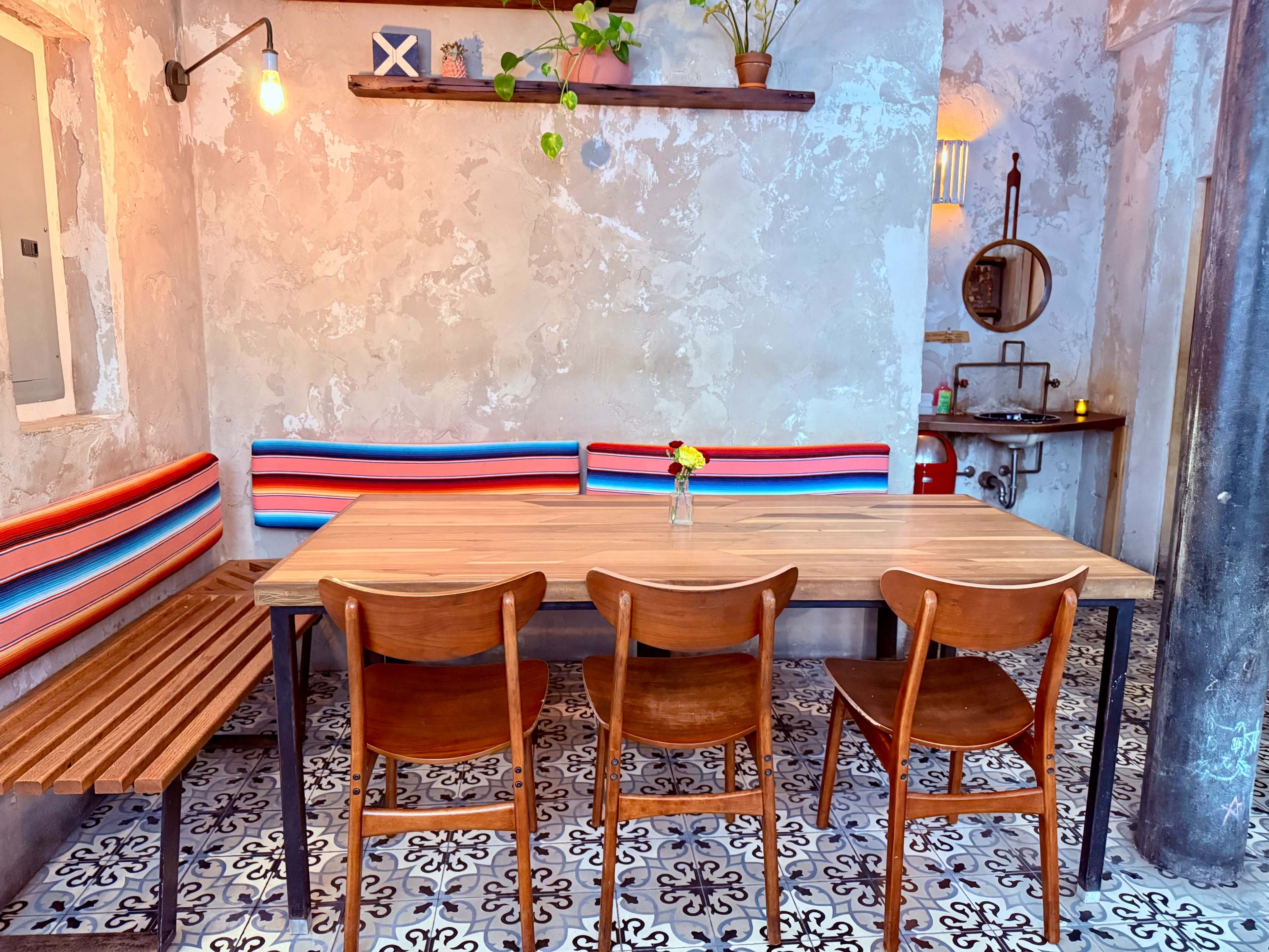 The image shows a cozy interior space with a wooden table surrounded by four wooden chairs, colorful striped benches along the walls, and decorative plants on a shelf.