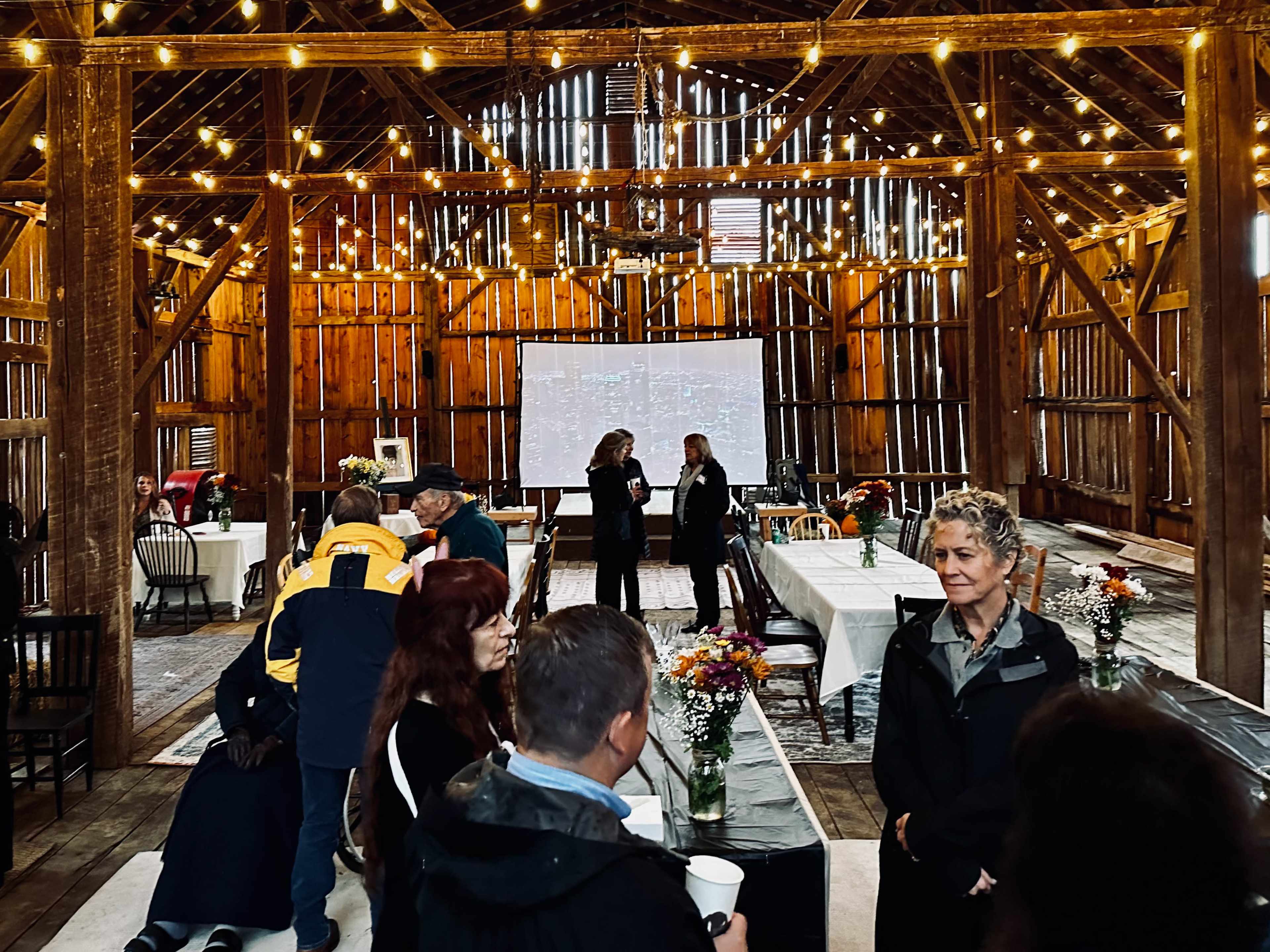 The interior of a rustic barn is set up for an event, featuring tables adorned with flowers and string lights, with attendees mingling throughout the space.