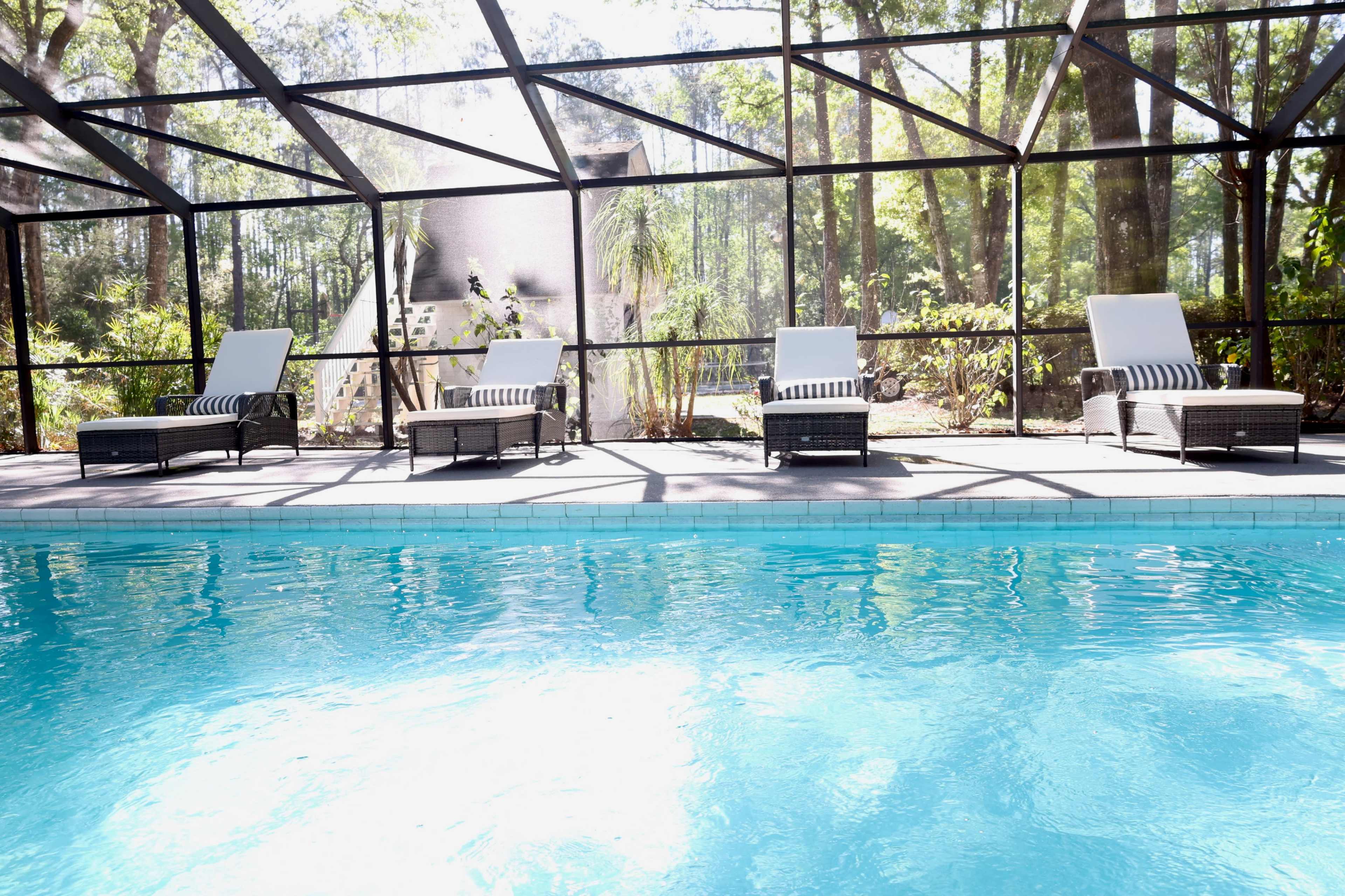 A row of four lounge chairs is positioned beside a clear blue pool within a screened patio surrounded by lush greenery.