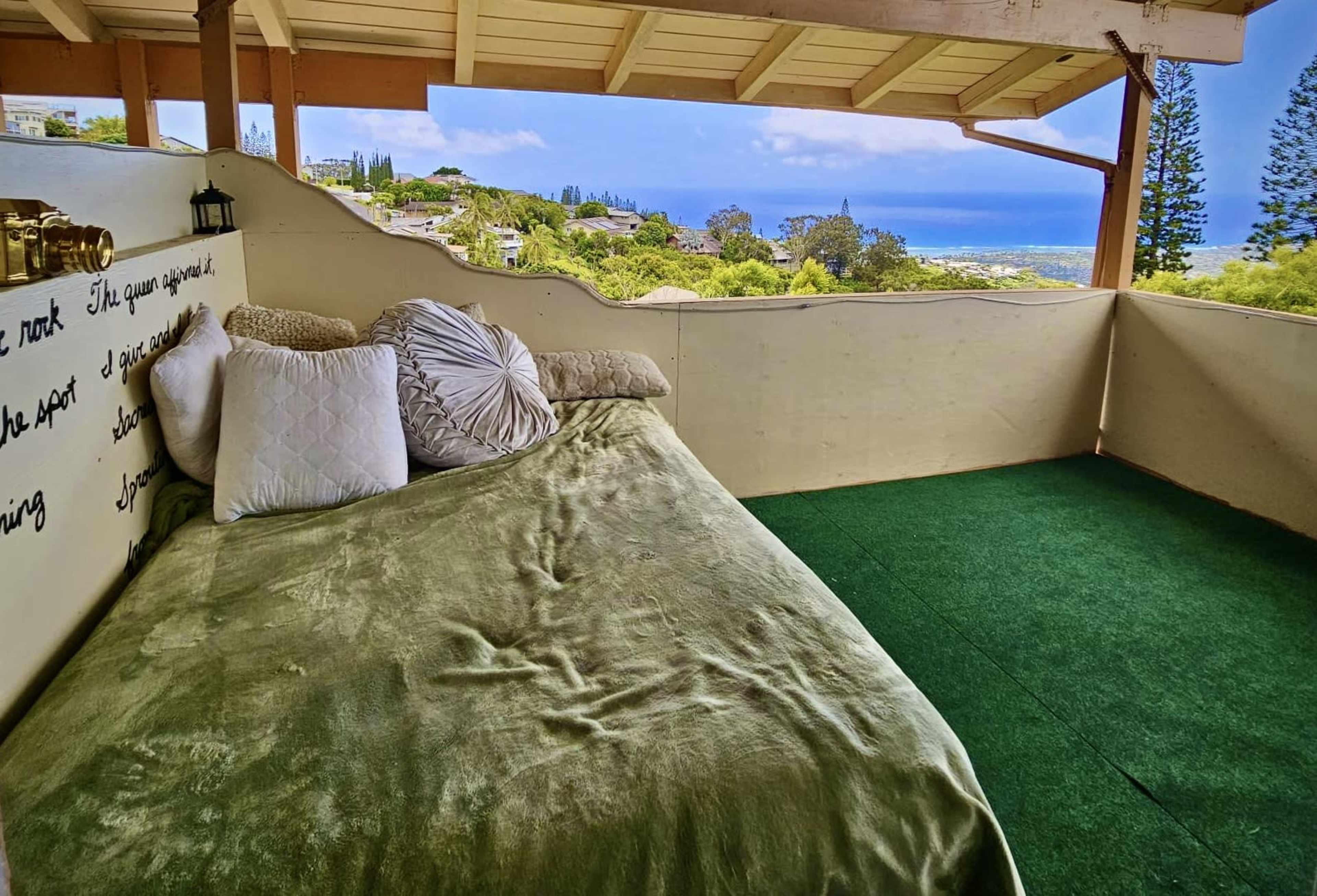 A cozy outdoor seating area with a plush green bed covered in pillows, overlooking a scenic view of greenery and the ocean in the distance.