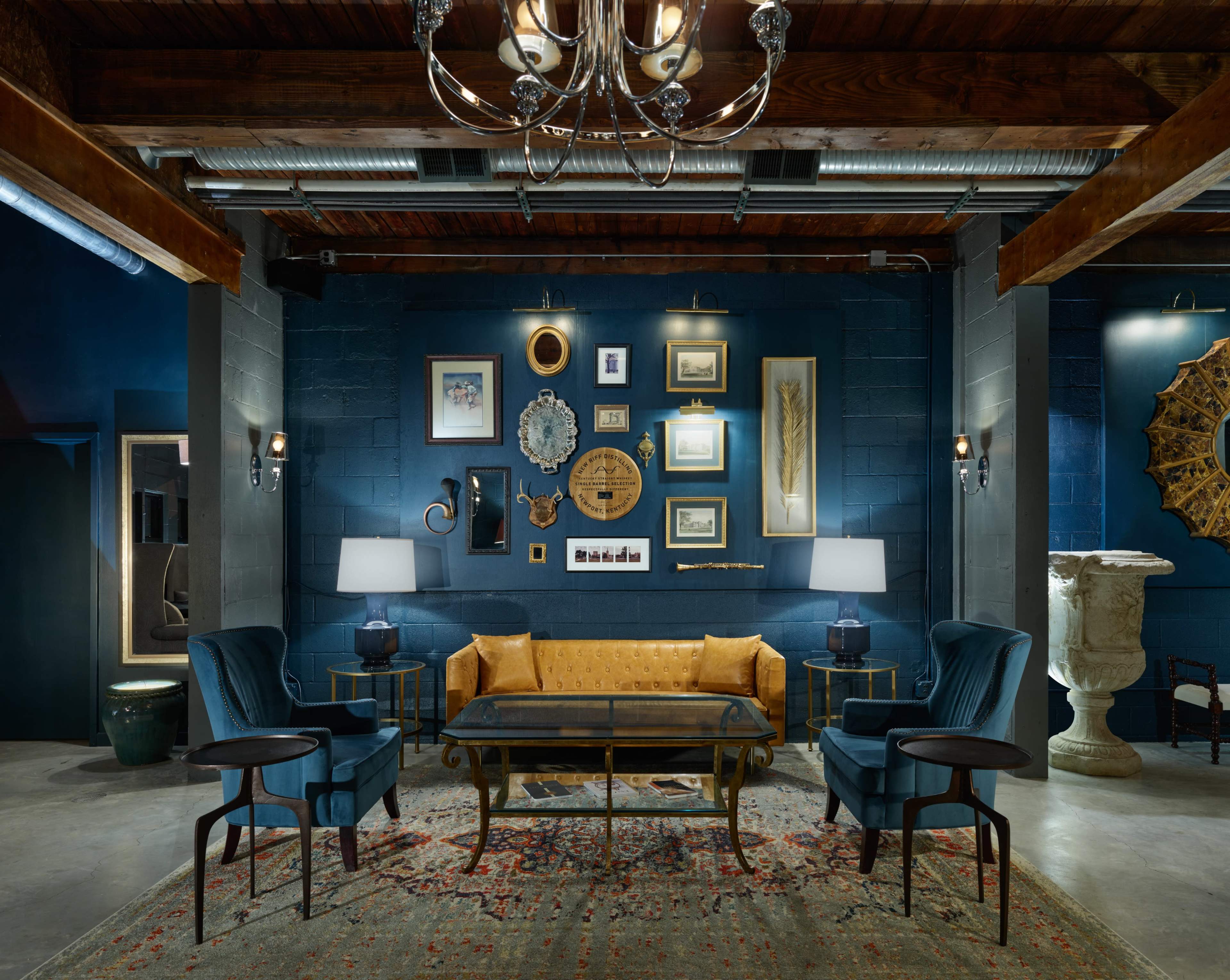 The image depicts a stylish lounge area with a blue wall adorned with framed artwork and decorative items, featuring a velvet couch, two blue armchairs, a glass coffee table, and elegant lighting fixtures.