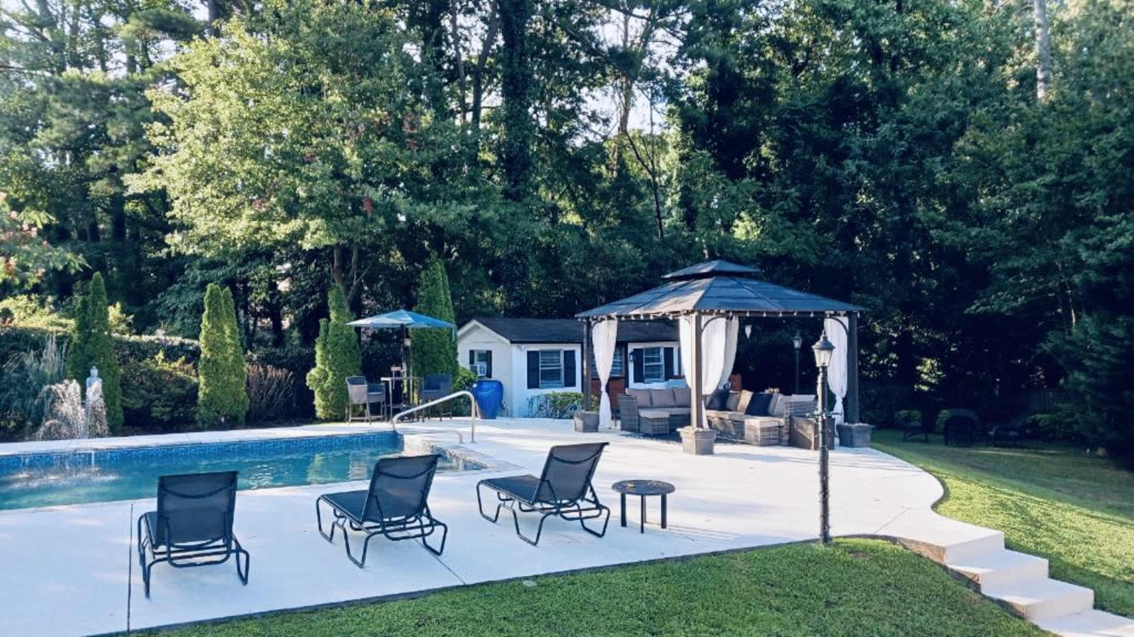 A backyard with a swimming pool, a gazebo, lounge chairs, and a well-maintained lawn surrounded by trees.