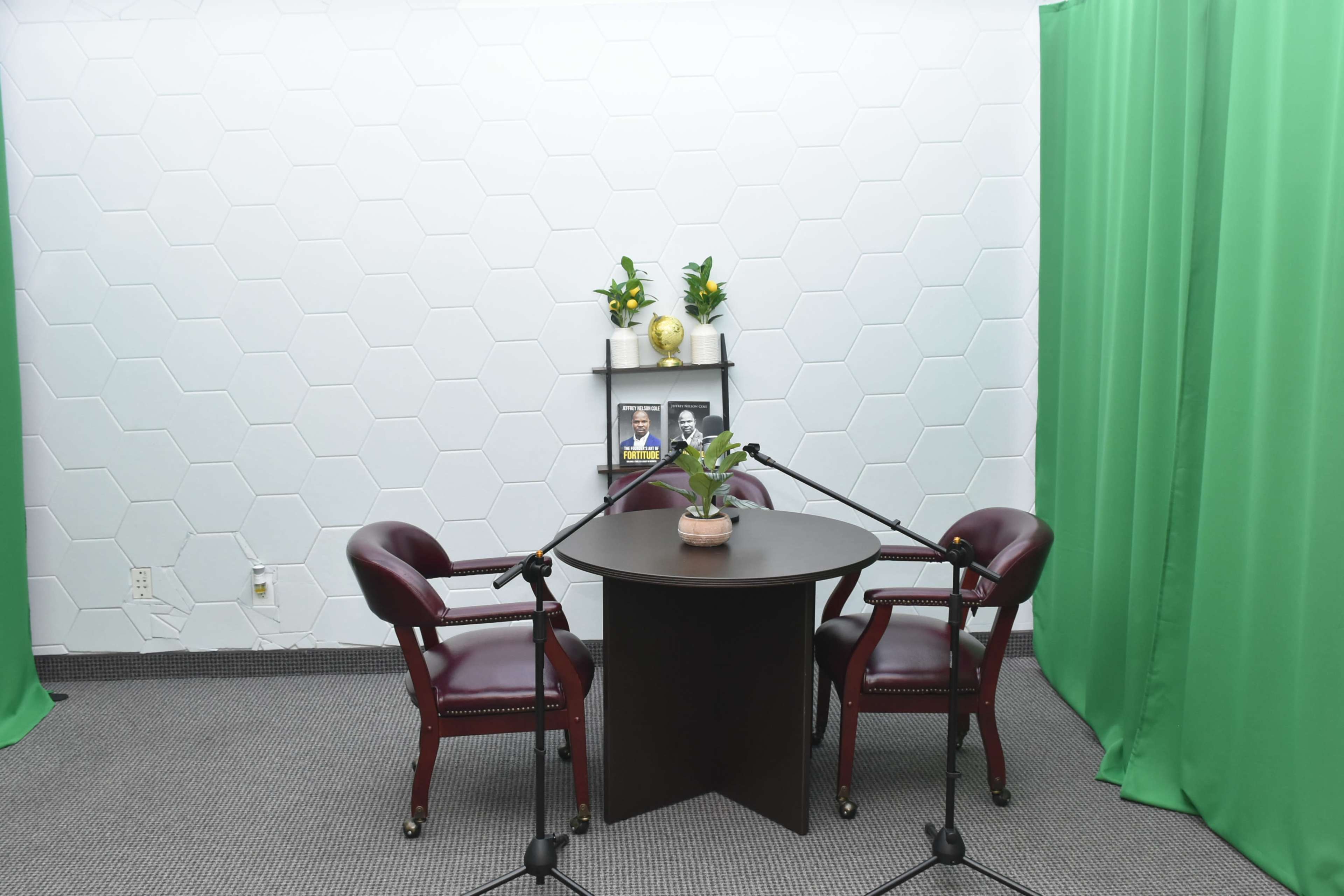 The image shows a small recording studio setup featuring a round table with two maroon chairs, flanked by green curtains and a shelf holding plants and framed pictures.