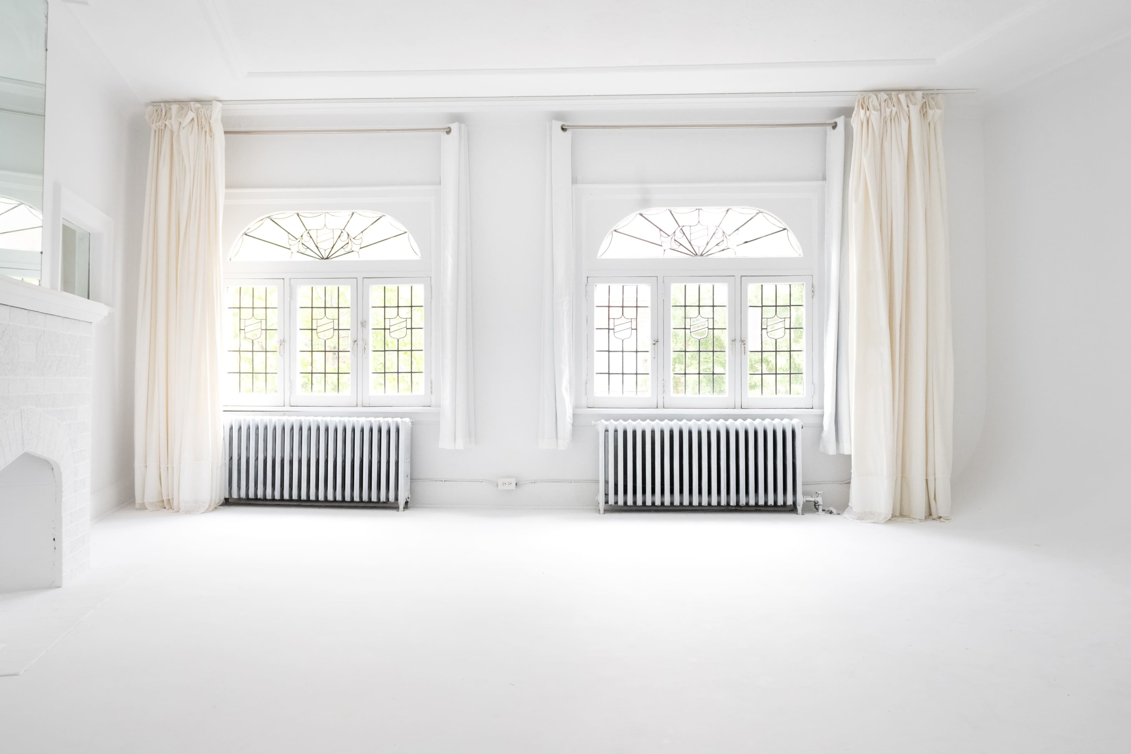 A bright, empty room with large windows and white walls, featuring two radiators below the windows.