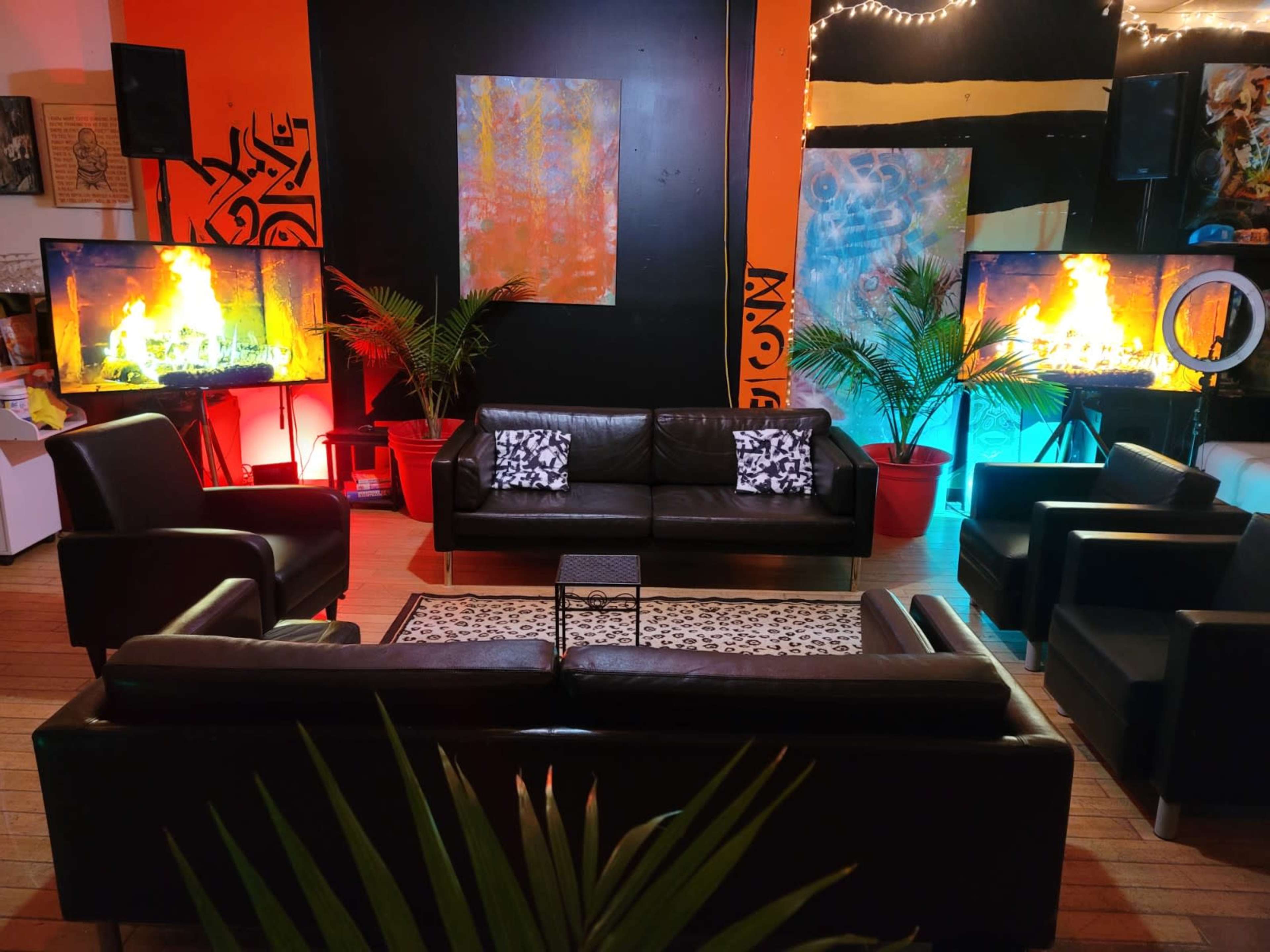 The scene features a modern lounge area with black leather furniture, two large televisions displaying flames, and colorful backgrounds illuminated by lights, complemented by potted plants.