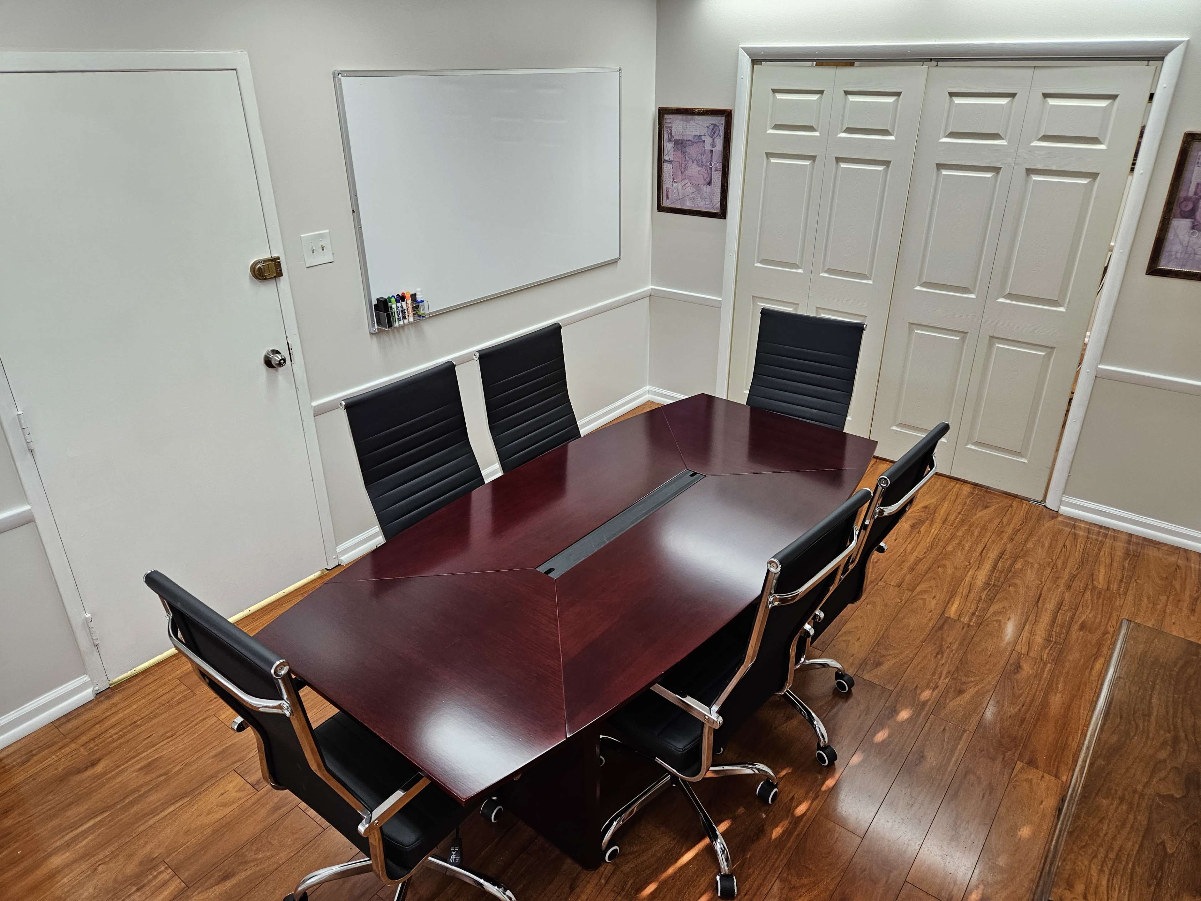 A conference room features a large, dark wooden table surrounded by six black office chairs, with a whiteboard and double doors in the background.