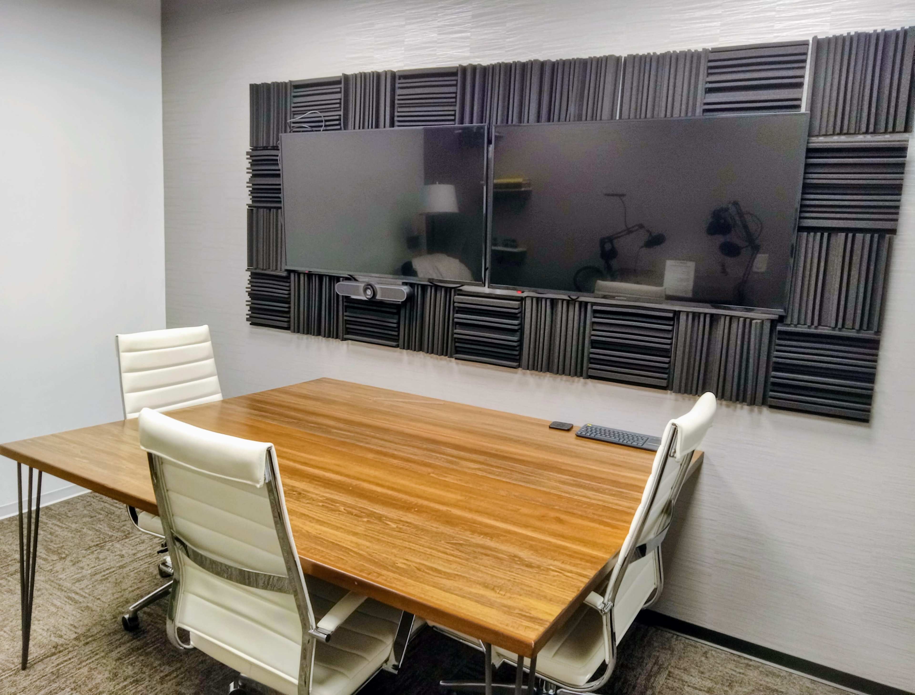 A modern conference room features a wooden table with white chairs and two large screens mounted on a wall with acoustic panels.