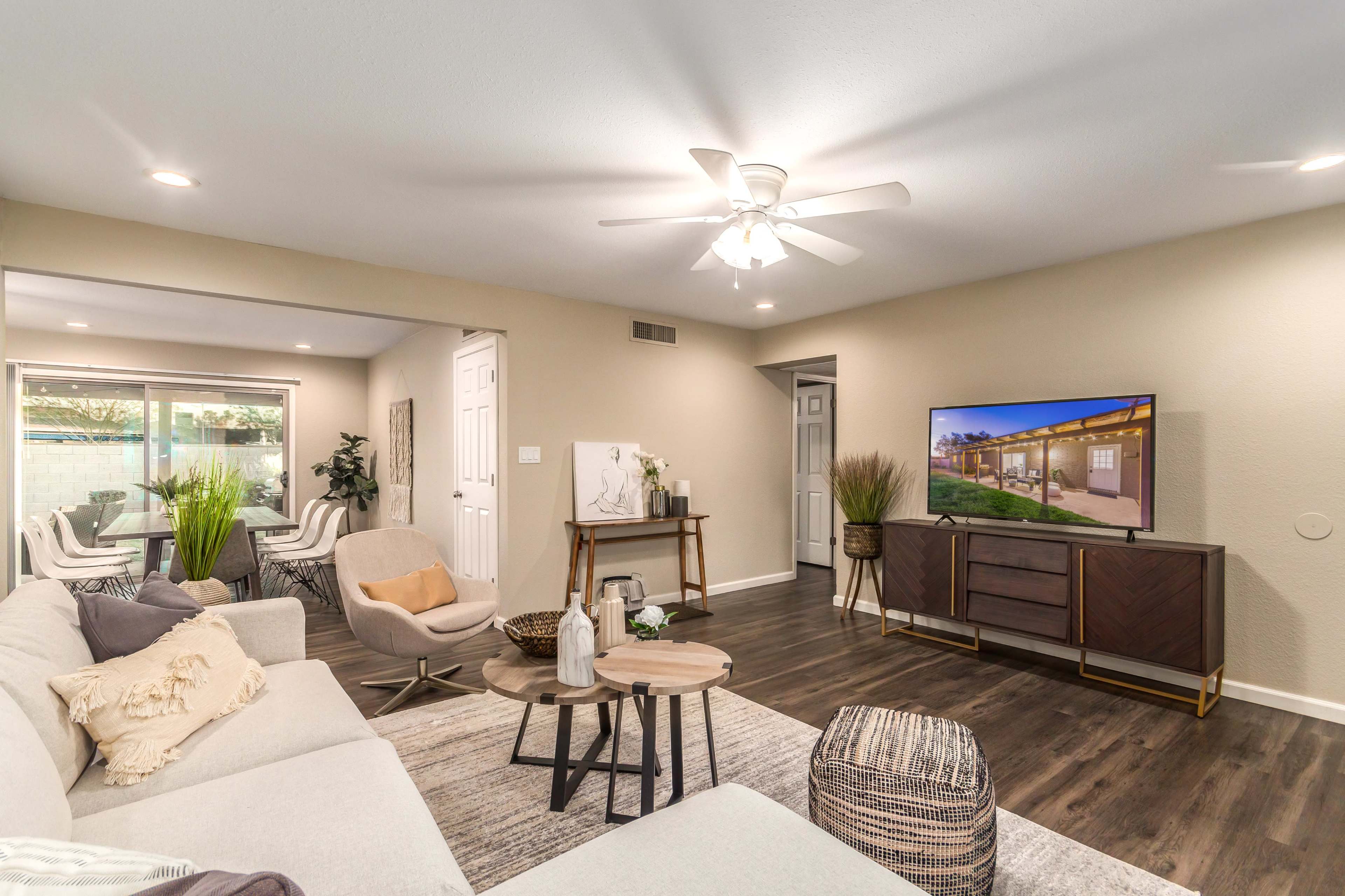 A modern living room features a beige sectional sofa, a coffee table, and a television on a stand, with decorative plants and bright lighting.
