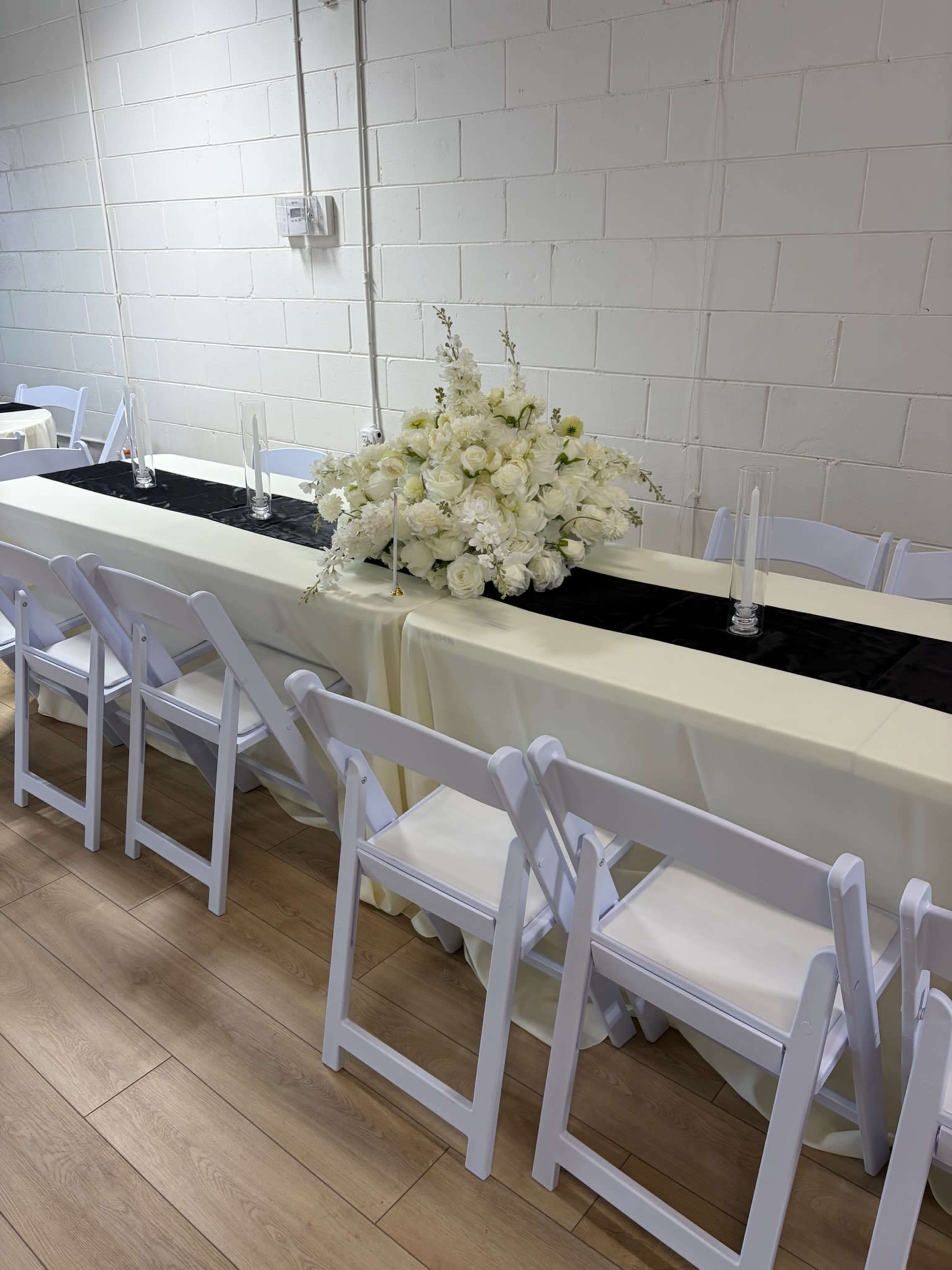 A long table is set with white flowers and a black runner, surrounded by white folding chairs in a well-lit room with exposed brick walls.
