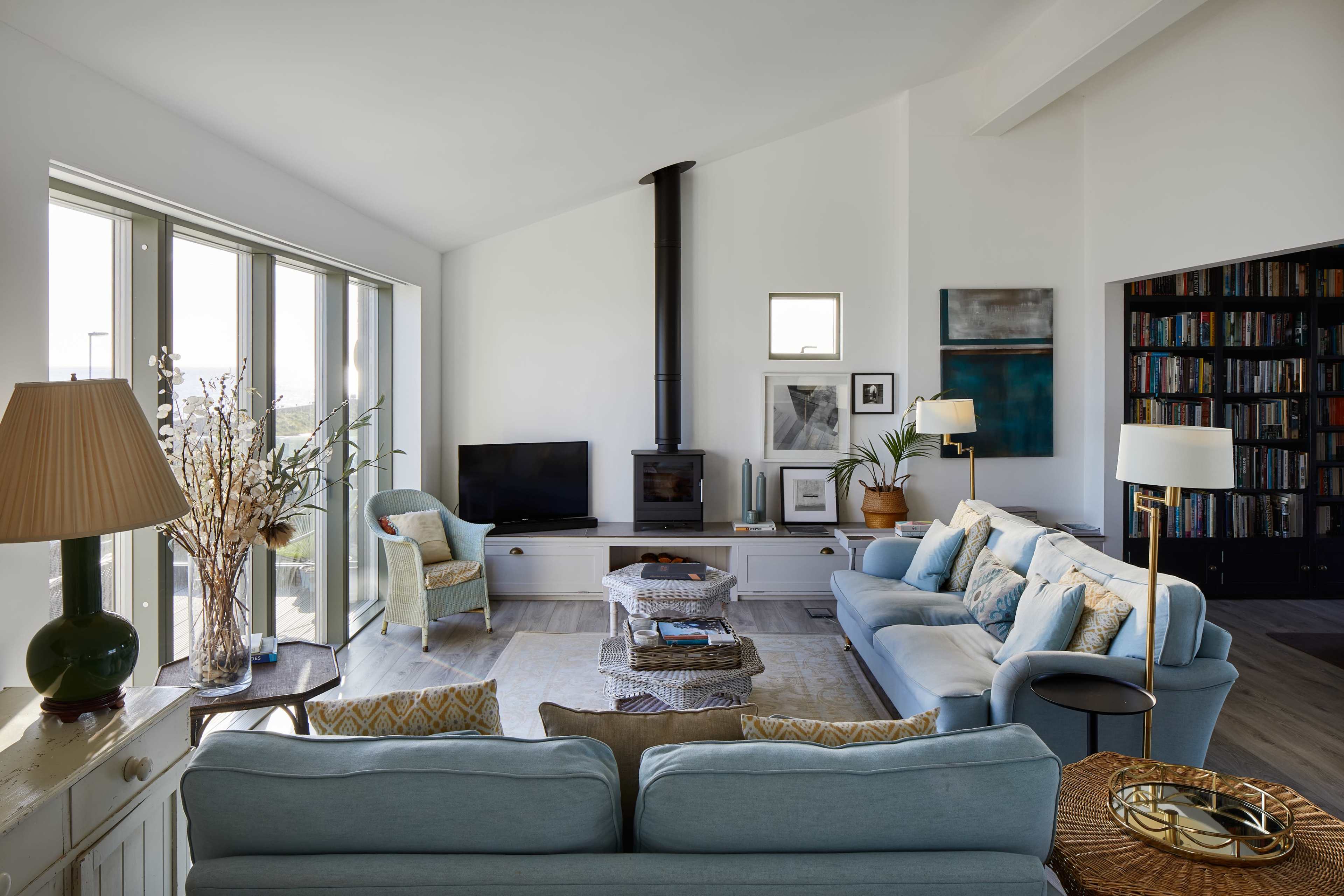 A modern living room features a light blue sectional sofa, an armchair, a wood-burning stove, and a large bookshelf, with ample natural light streaming through tall windows.