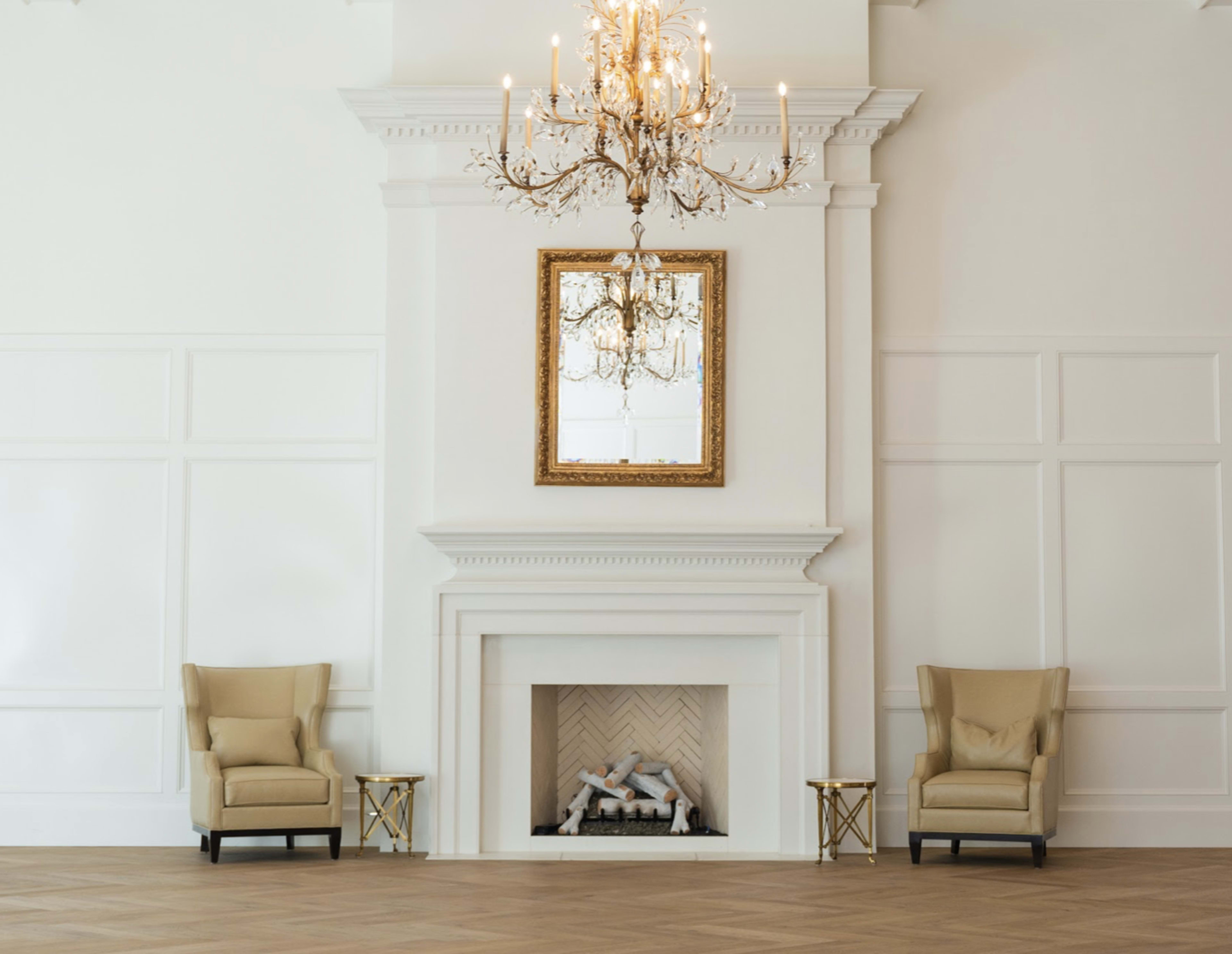 The image shows a spacious room featuring a white fireplace with a herringbone pattern, flanked by two beige armchairs and a chandelier overhead.