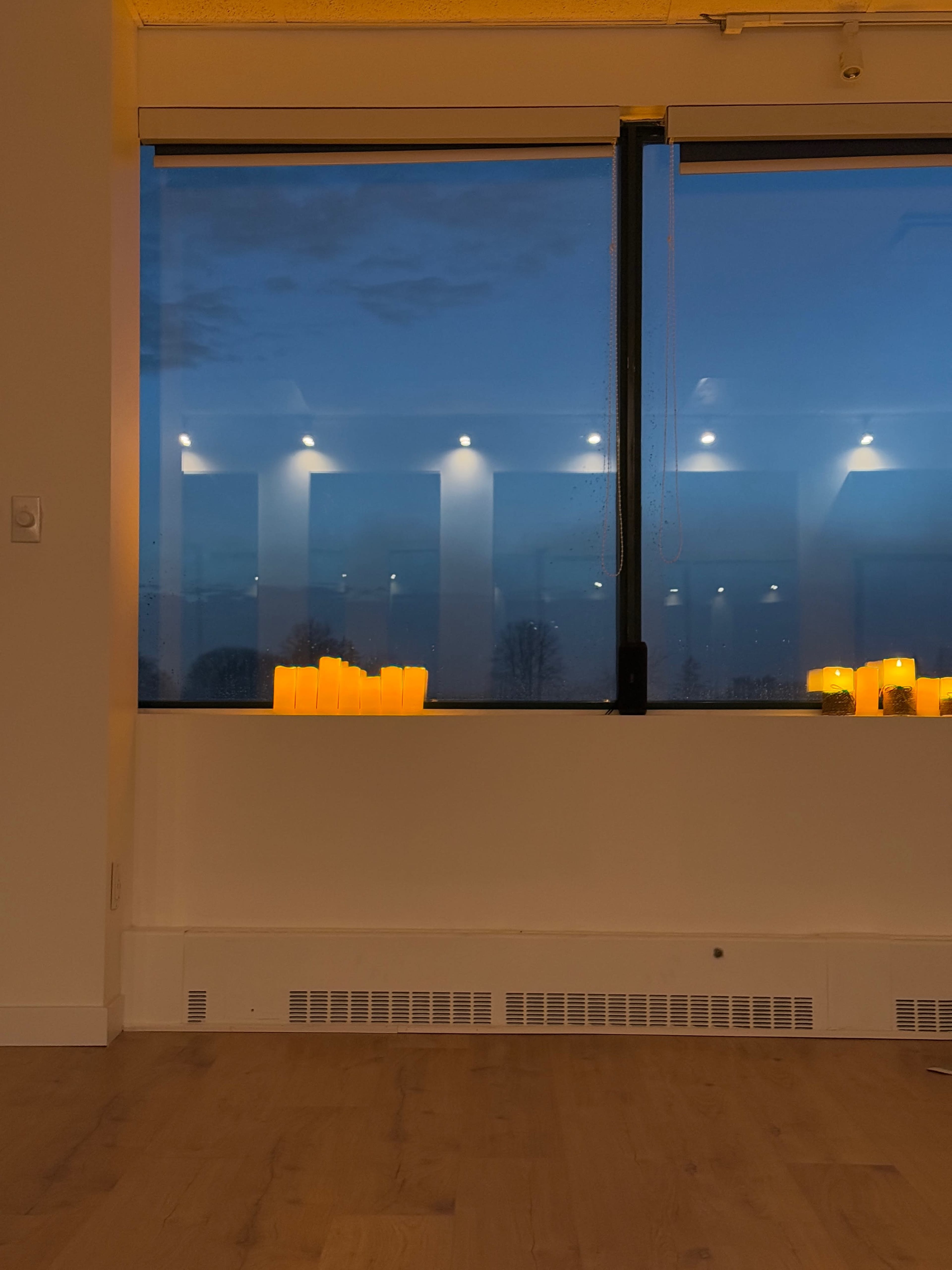 The image shows a window with a view of a dimly lit room featuring flickering candles arranged on a ledge against a dusk sky.