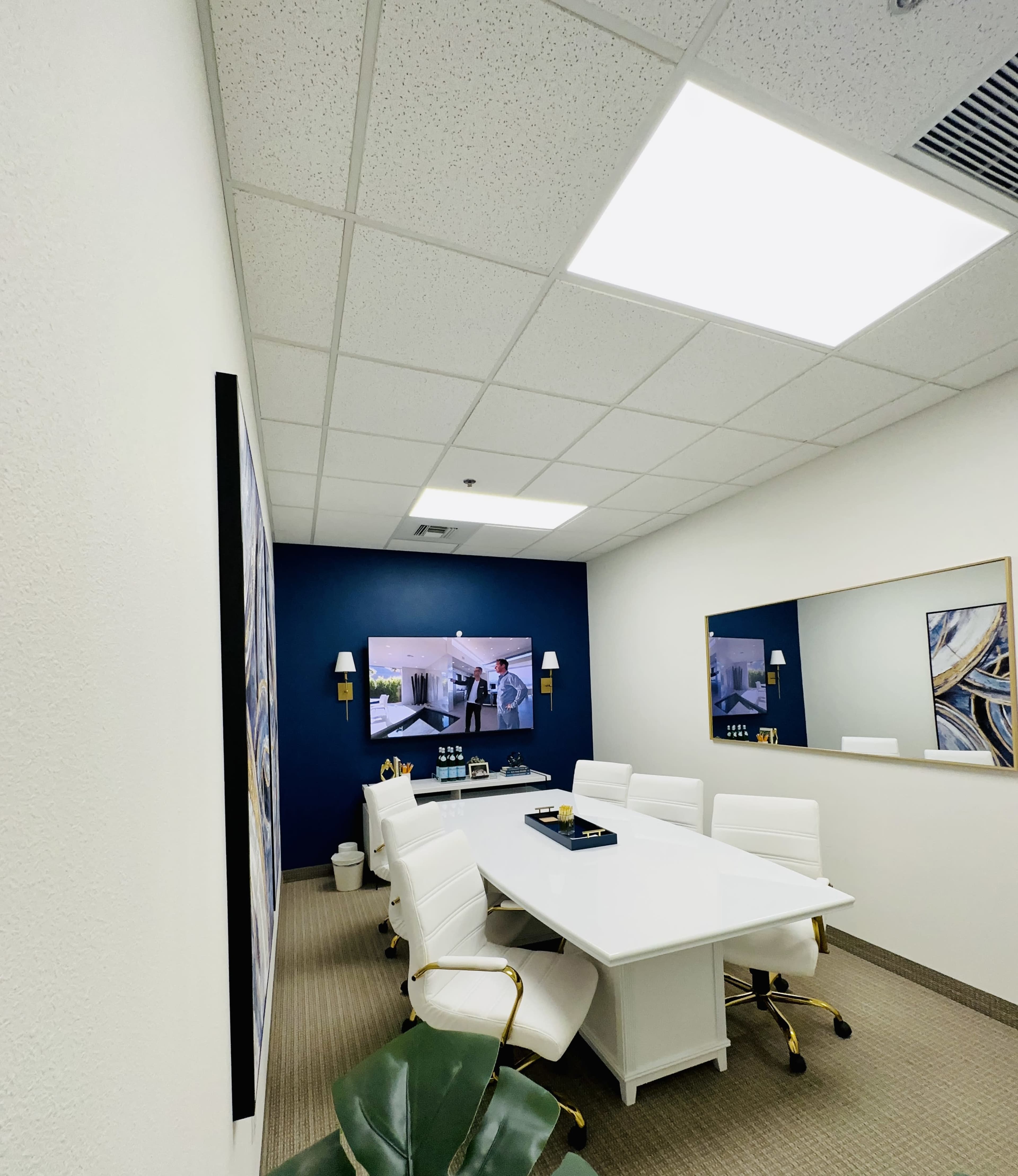 A modern conference room features a large white table surrounded by white chairs, with a blue accent wall and two televisions displaying images.