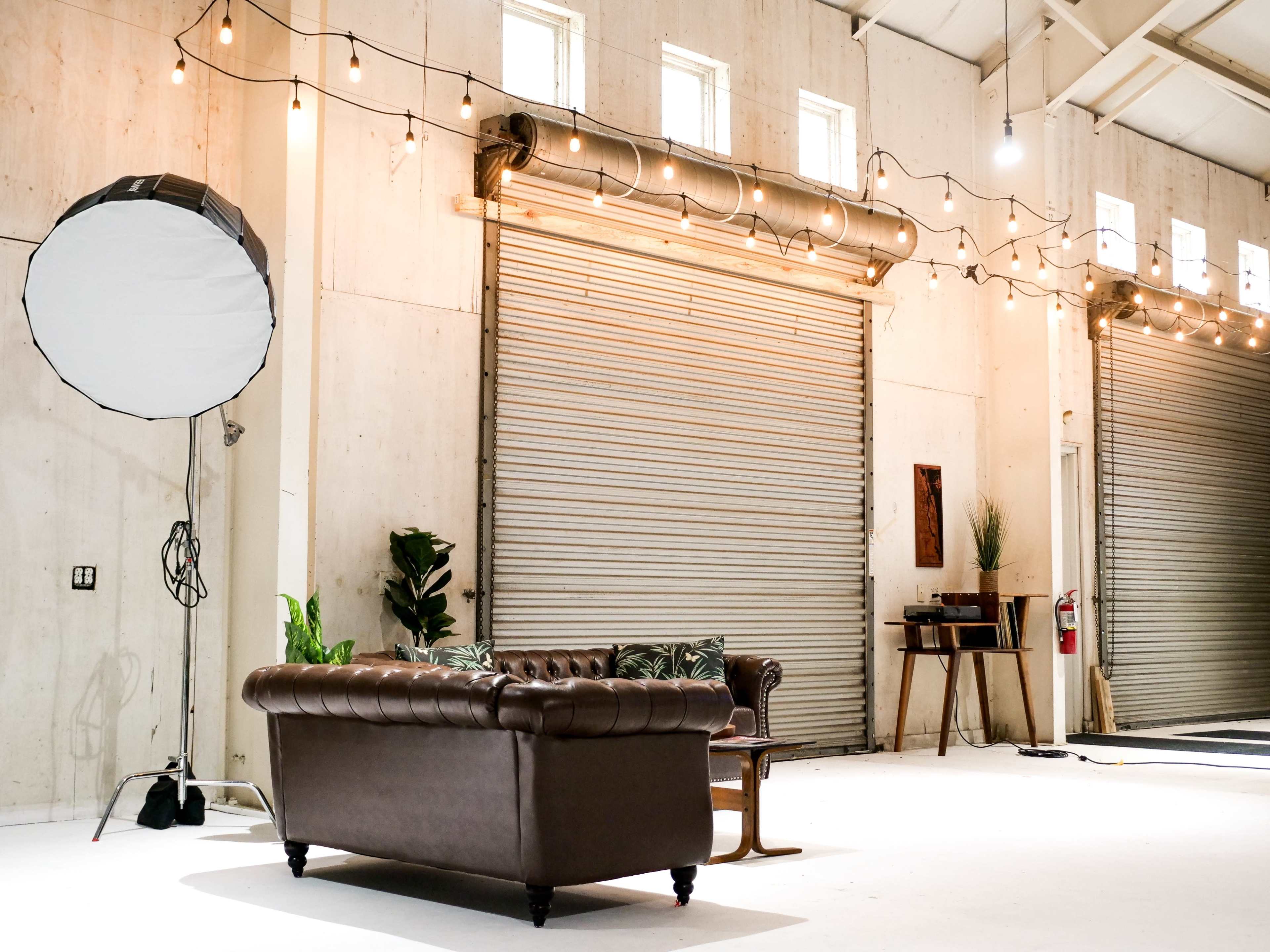 A spacious interior with a brown leather couch, a photography light setup, and decorative string lights against a backdrop of closed roll-up doors.