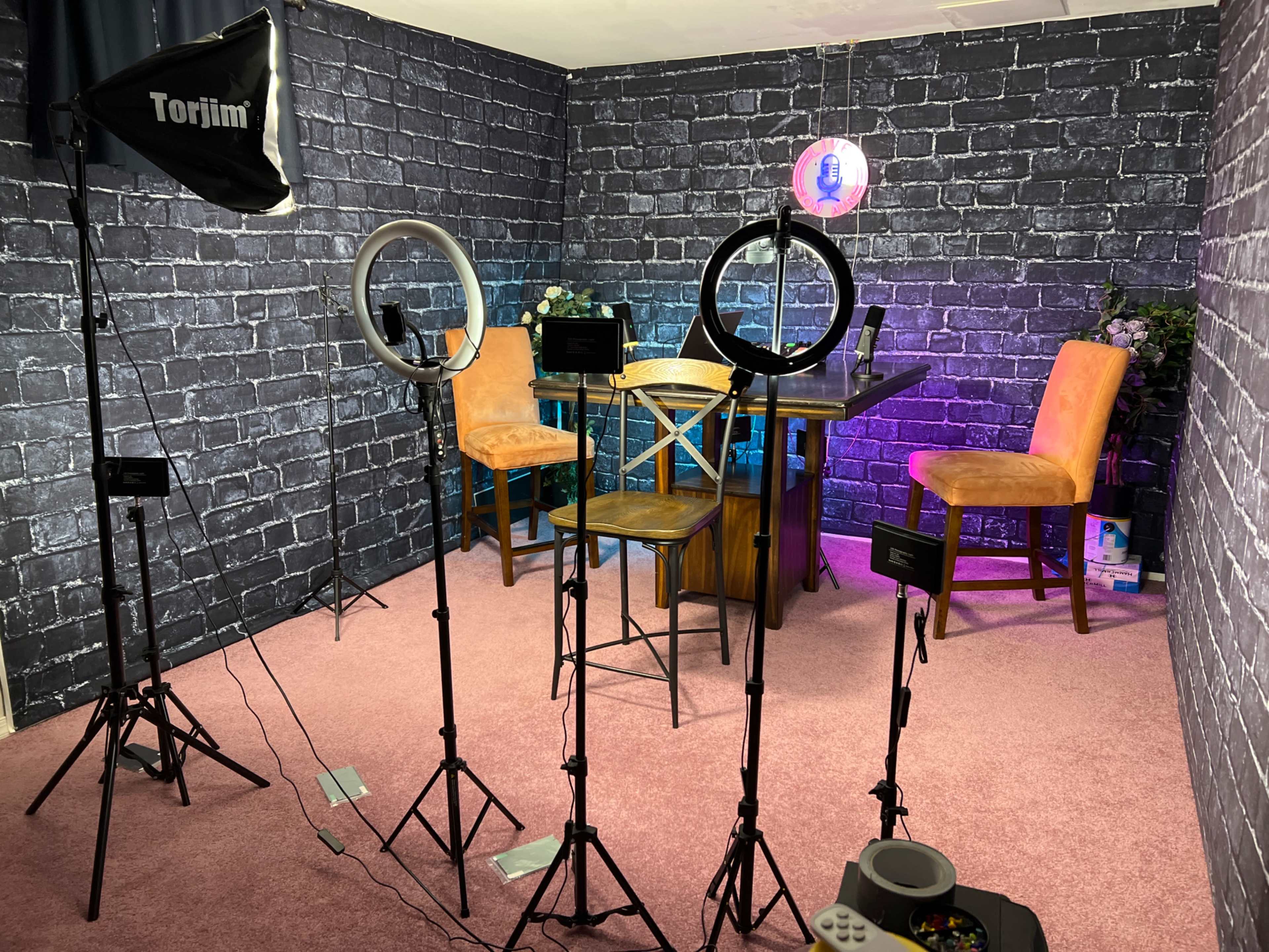 A studio setup with lighting equipment and a wooden table surrounded by chairs against a brick-patterned wall.