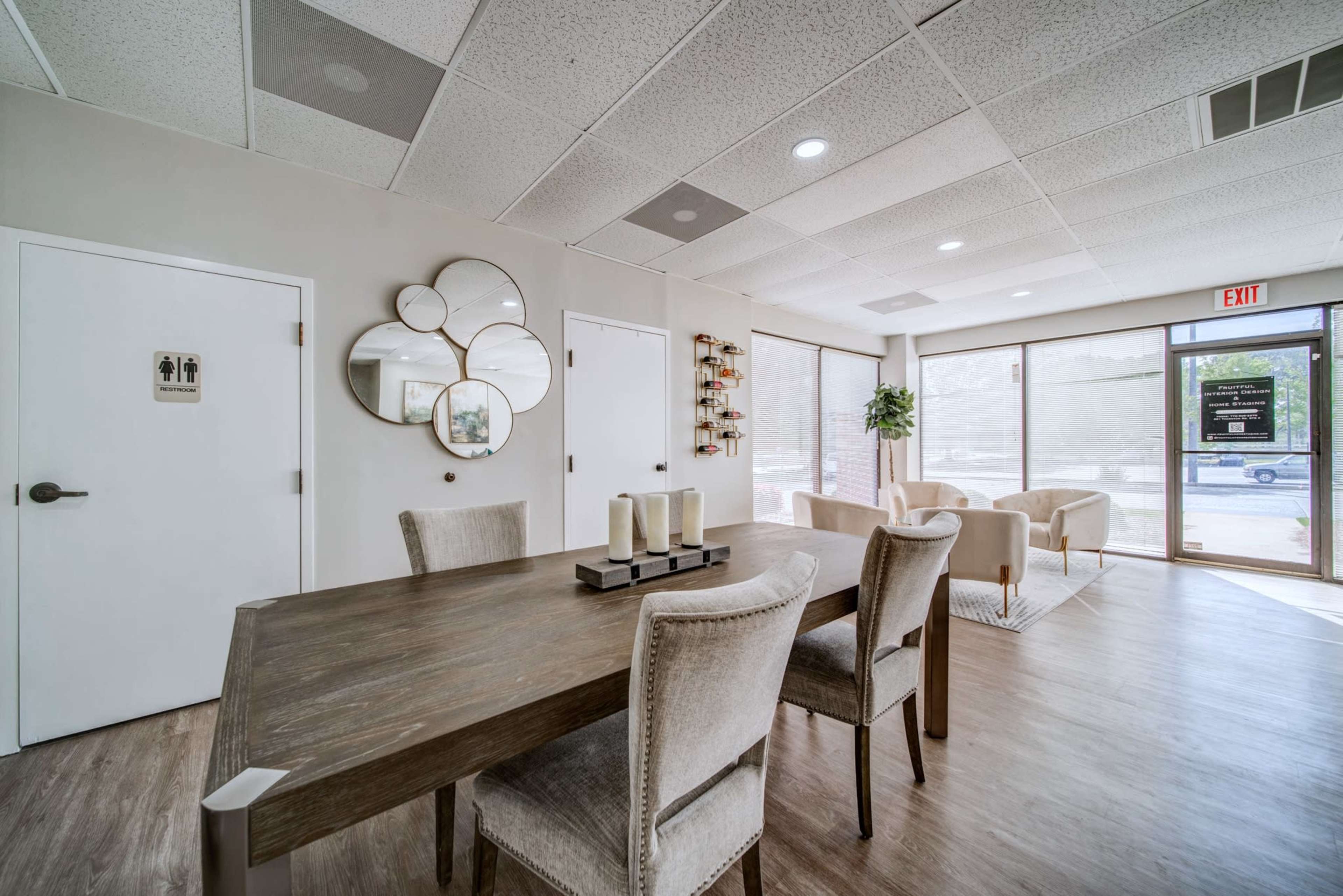 The image shows a modern waiting area with a dining table, upholstered chairs, a round mirror on the wall, and large windows letting in natural light.