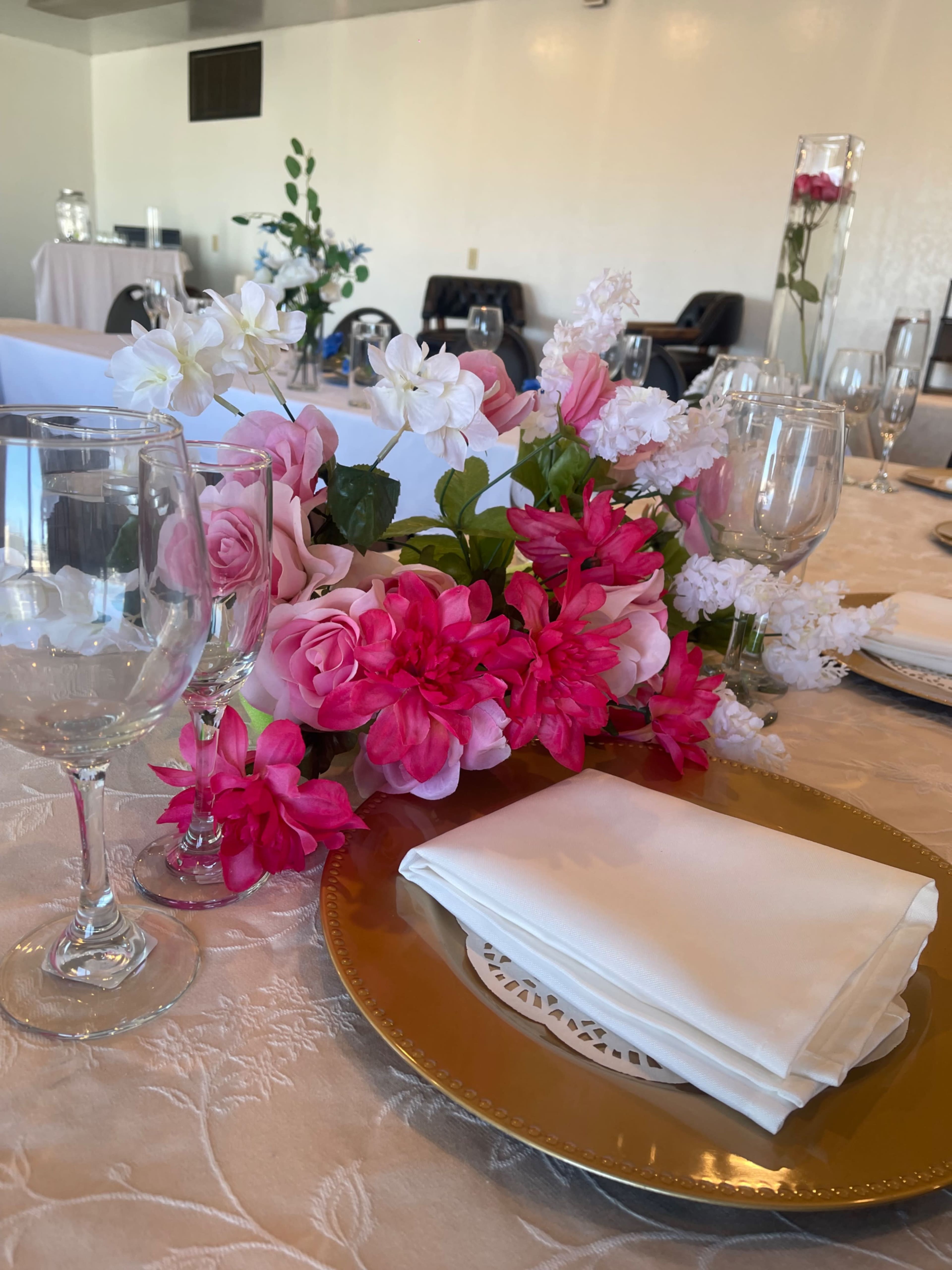 A table is set with white napkins on gold plates, surrounded by an arrangement of pink and white flowers and glassware.