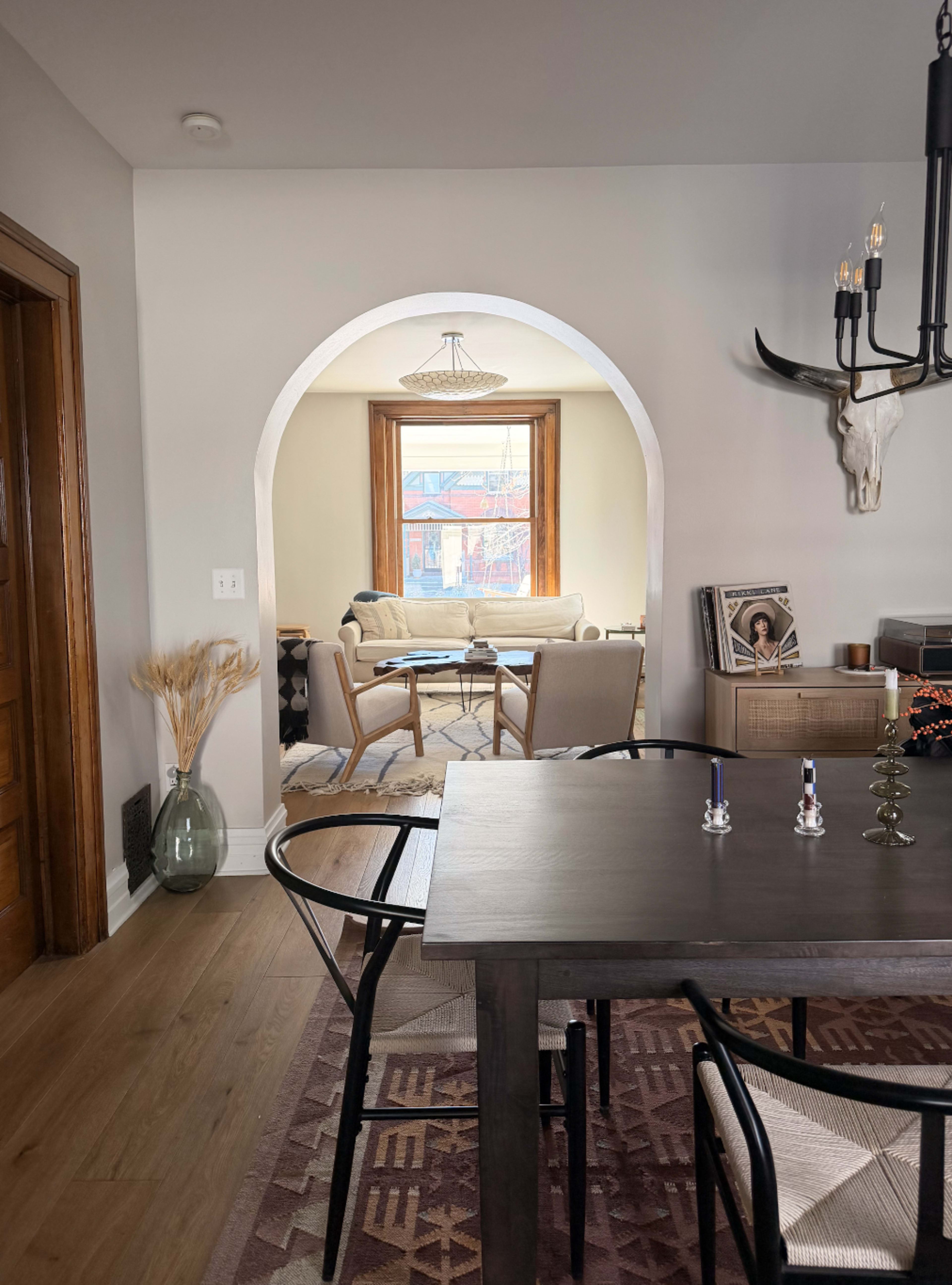 The image shows a dining area with a table and chairs, leading to a living space with a window and a couch beyond an arched doorway.