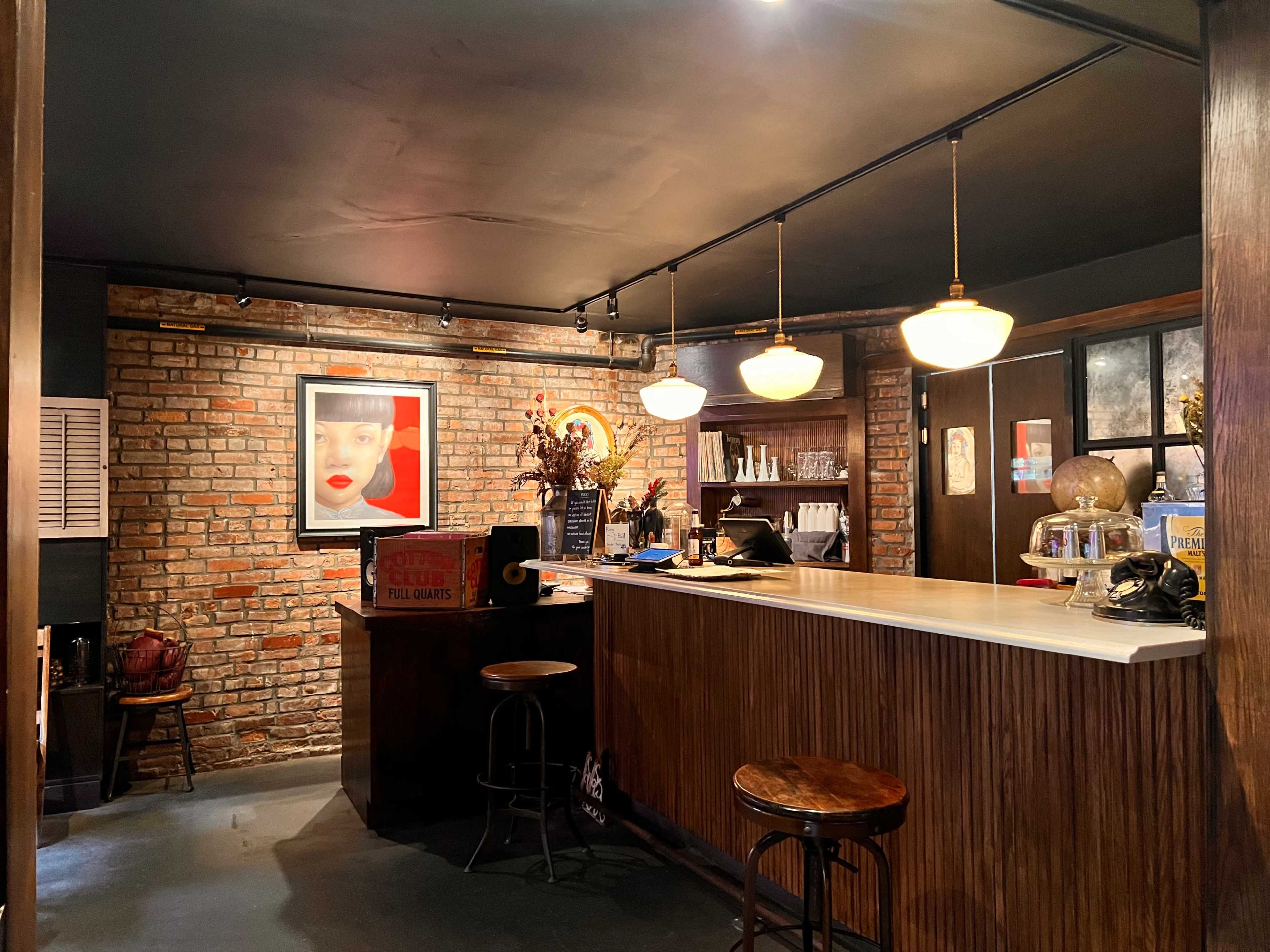 The image shows a dimly lit bar area with a brick wall, a wooden counter, and two bar stools, adorned with various decorative elements.