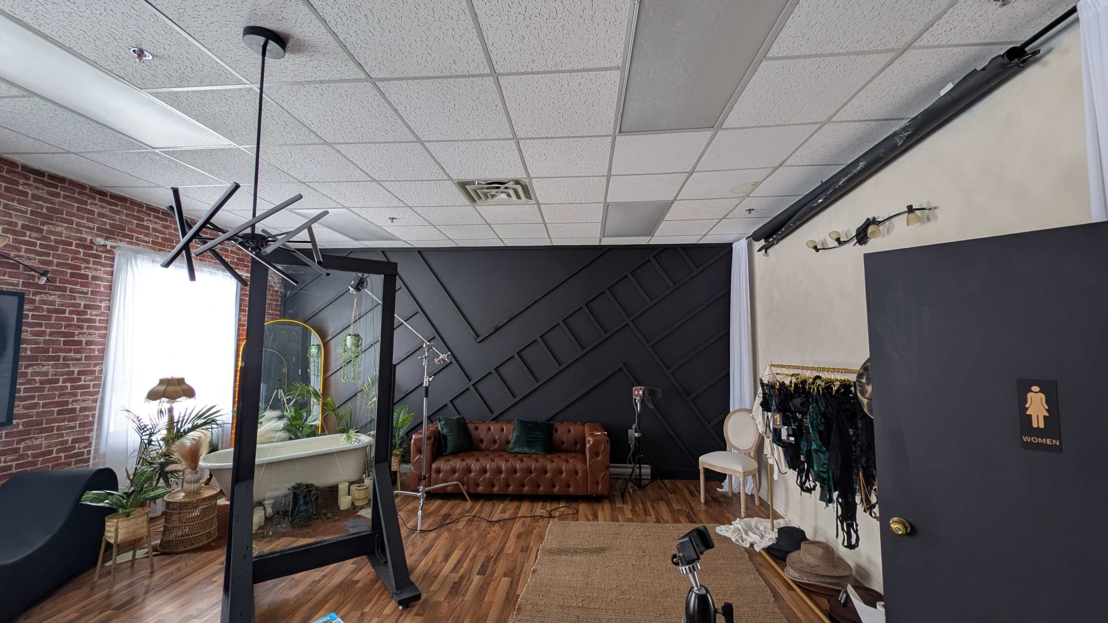 The image shows a modern studio space featuring a leather sofa, a makeup mirror, clothing racks, and a backdrop on opposite walls, with wooden flooring throughout.