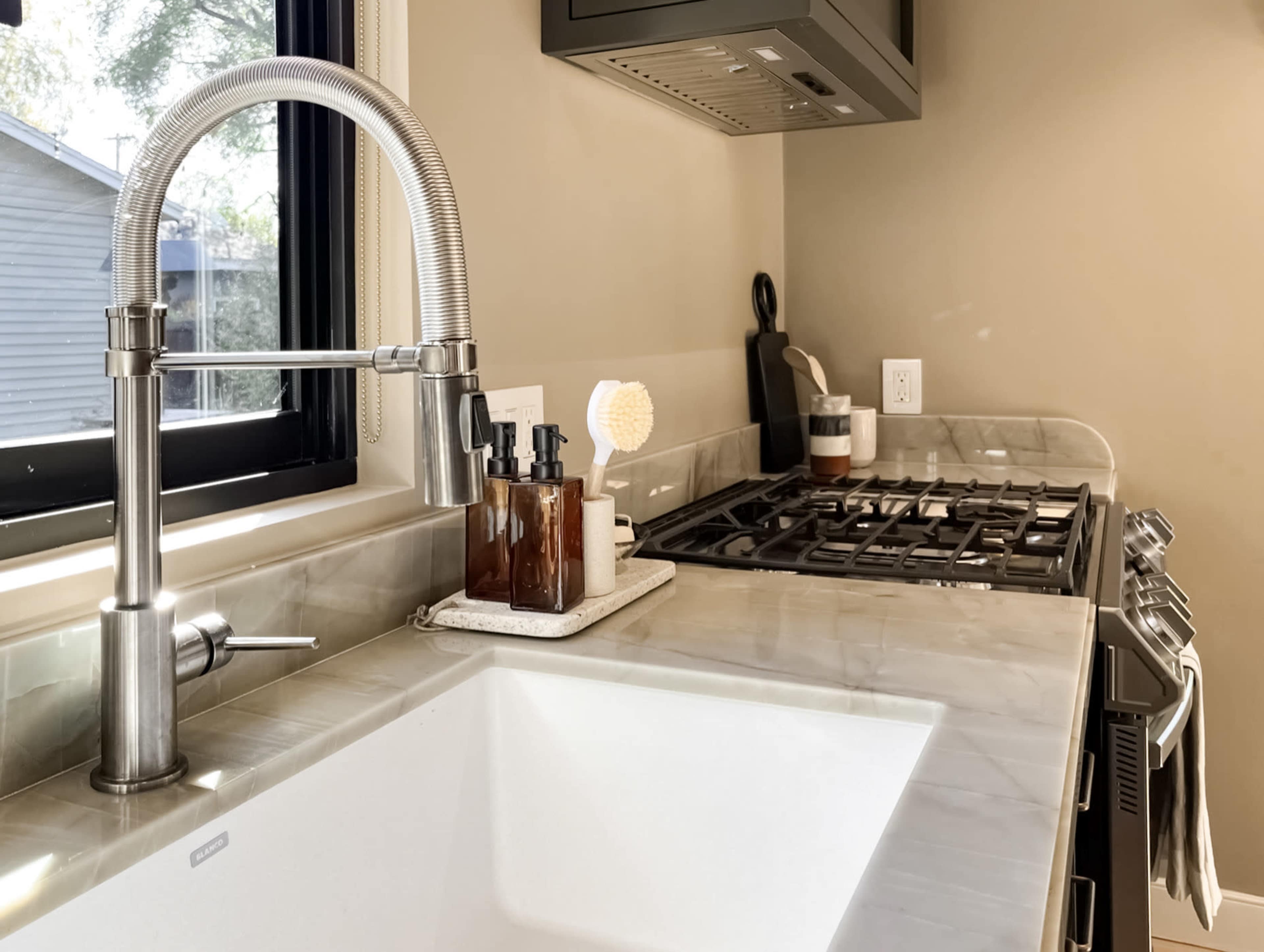 A modern kitchen scene features a stainless steel faucet above a large white sink, with a gas stove and various kitchen items organized on the counter.