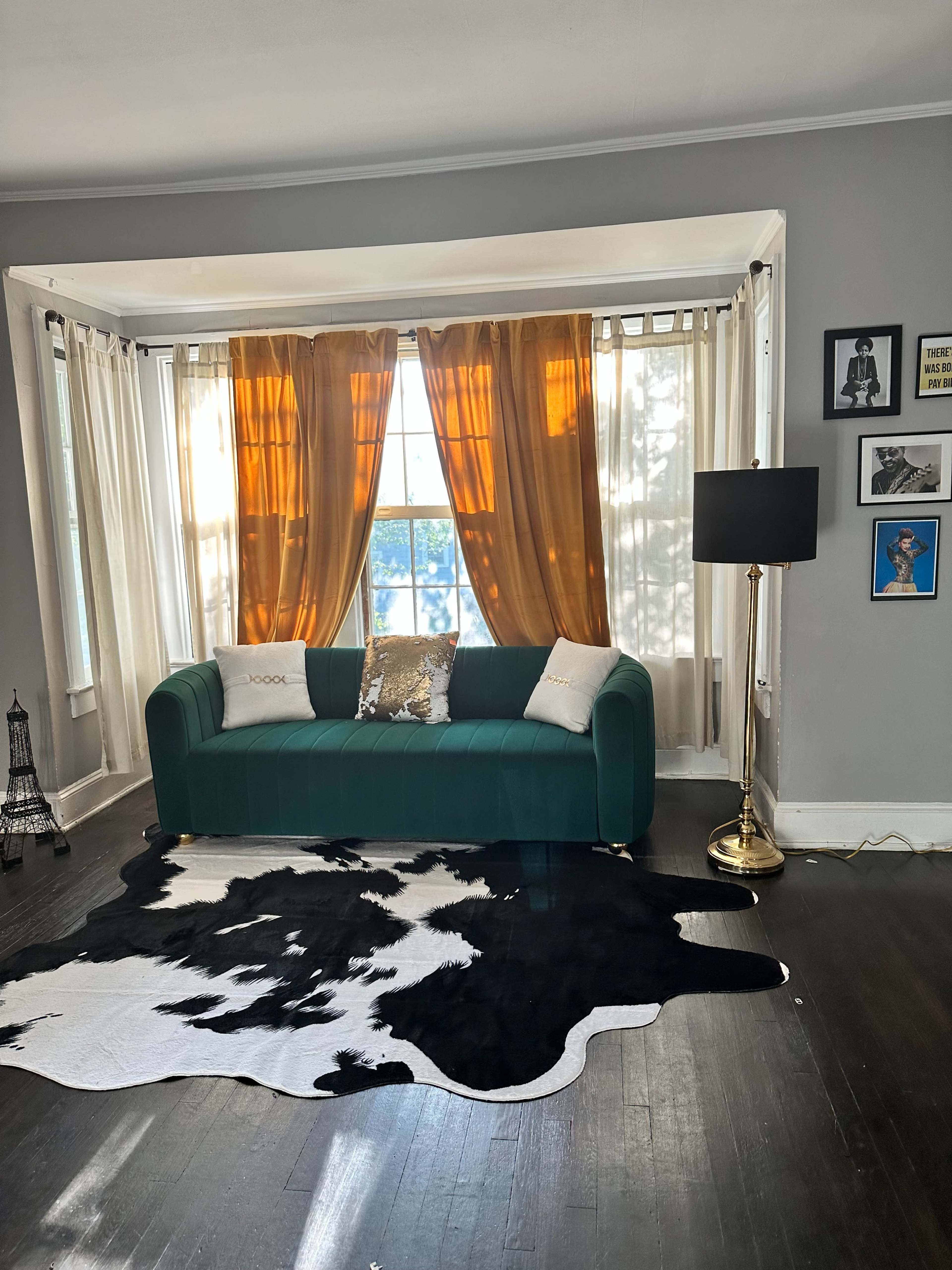 A green couch with a gold pillow is positioned in front of a large window adorned with orange curtains, alongside a black and white cowhide rug and a standing lamp.