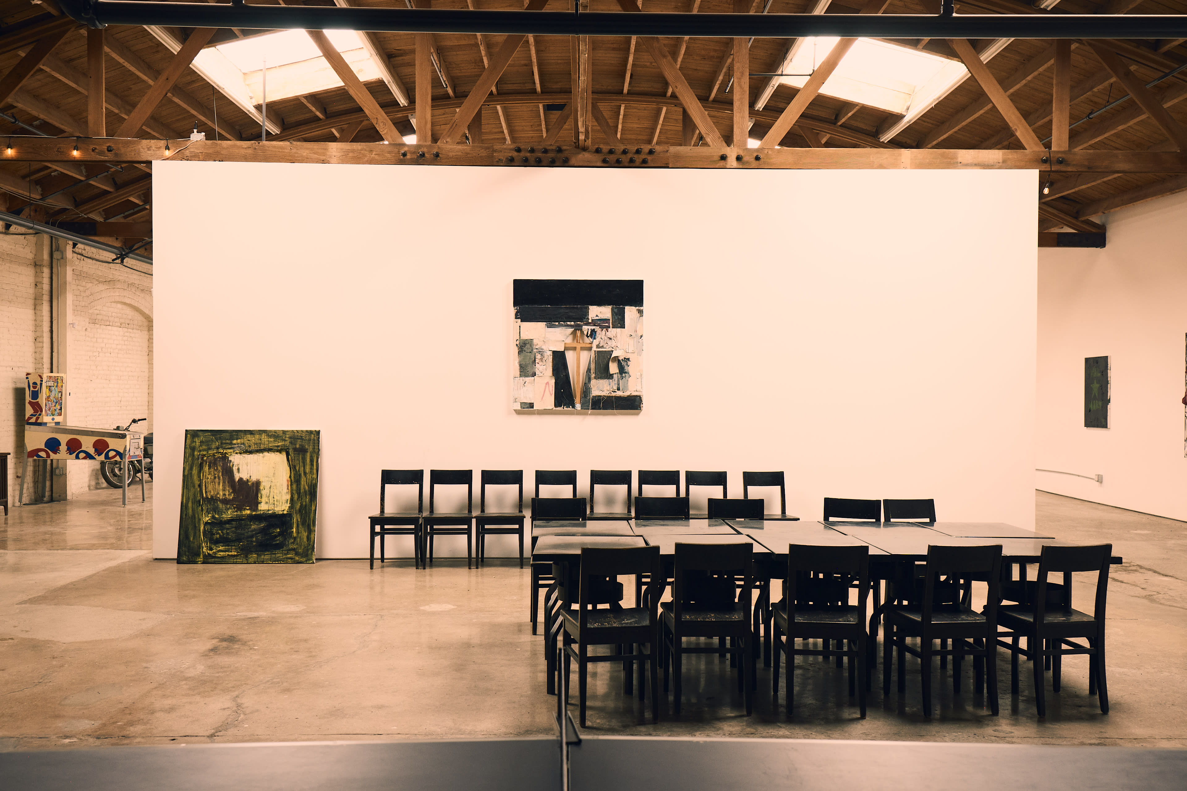 The image shows a spacious art gallery featuring a large, empty table with chairs arranged around it, a painting on the wall, and an artwork leaning against the side.