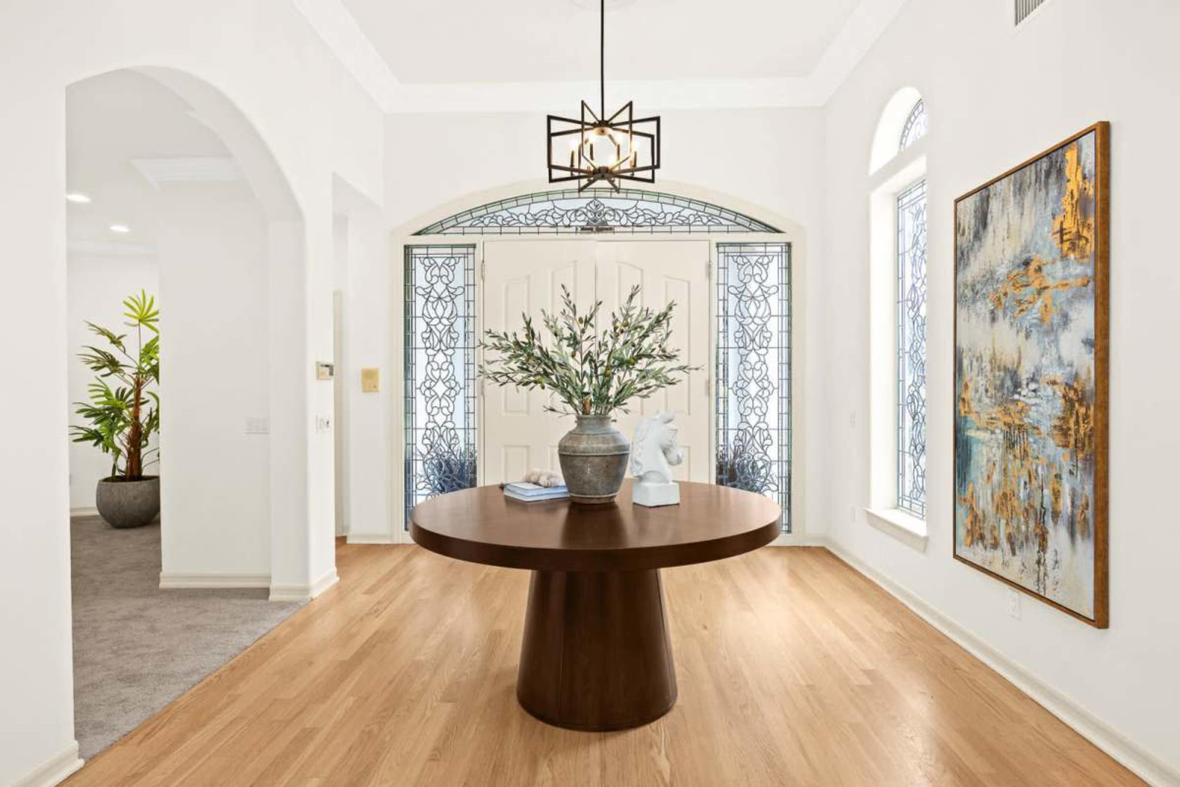The image shows a modern entryway featuring a round wooden table at the center, flanked by large decorative windows and a piece of abstract art on the wall.