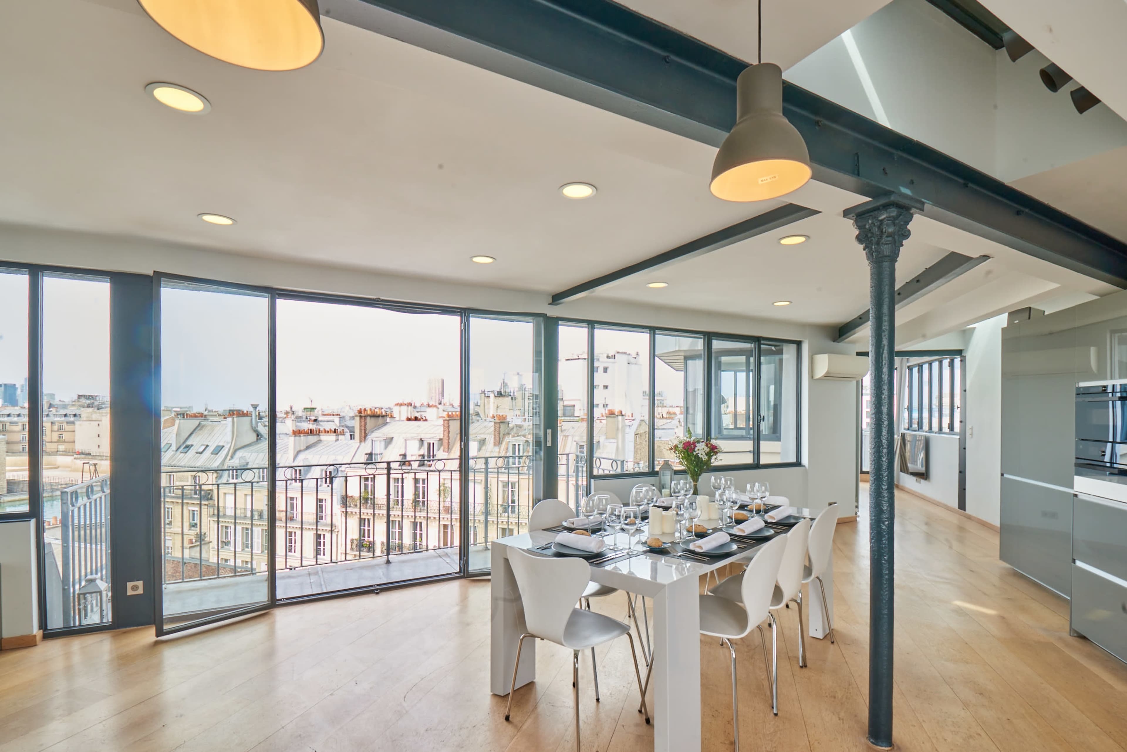 A modern dining area with a large table set for a meal, surrounded by large windows offering a view of the rooftops and skyline outside.