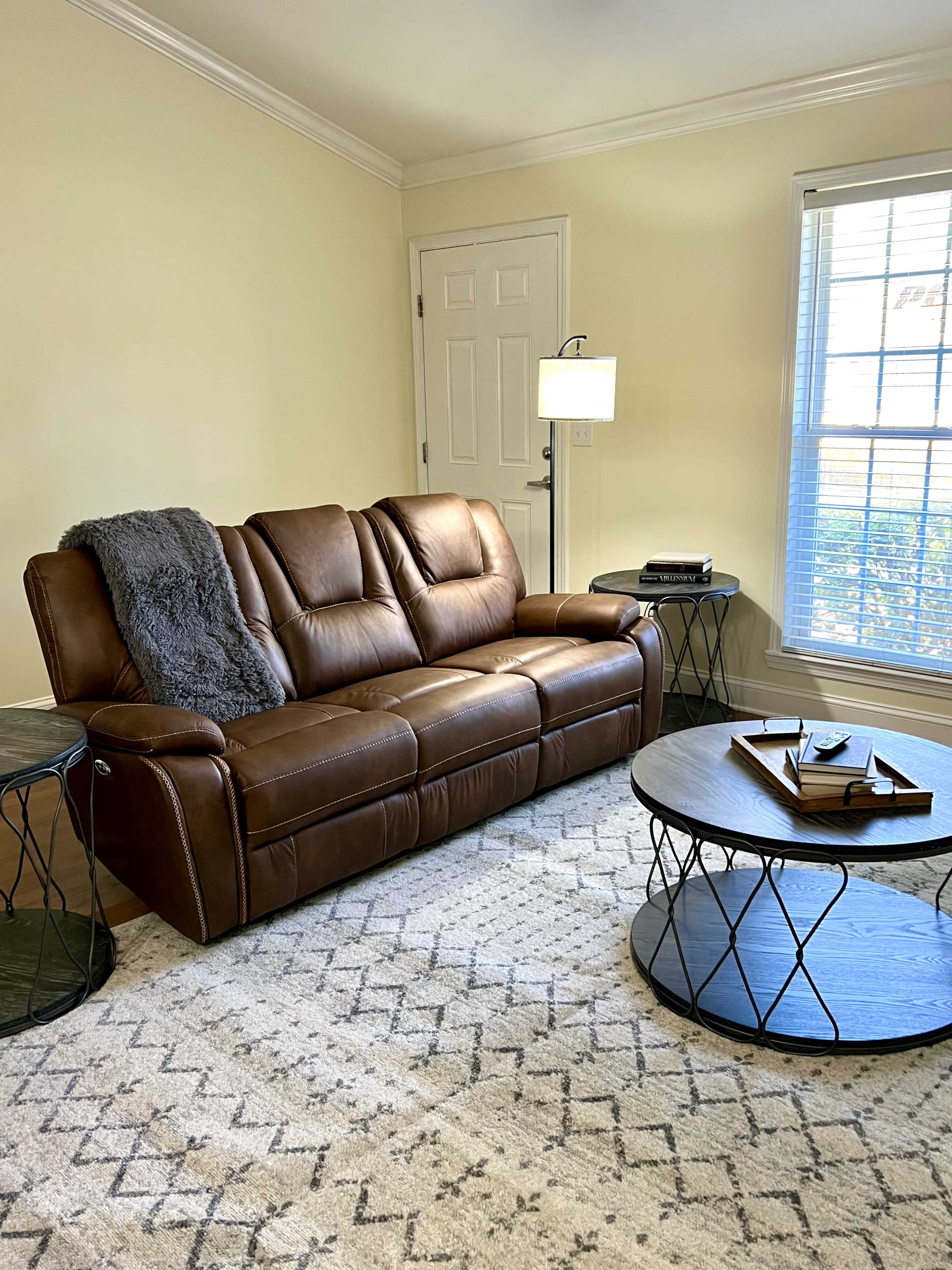A brown leather couch is positioned in a well-lit room with two black wireframe tables and a window featuring white blinds.
