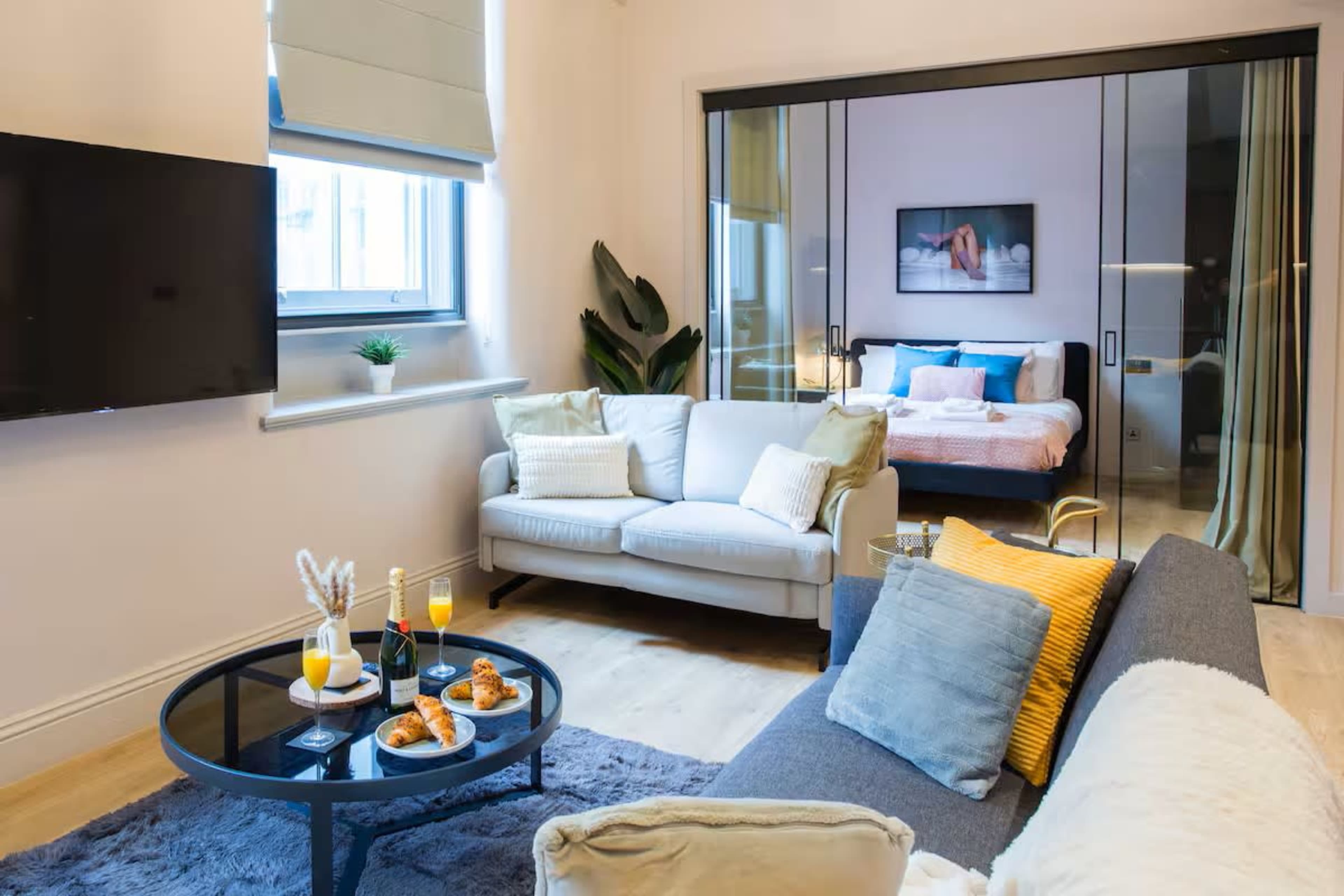 The image shows a modern living space featuring a gray sofa, a round coffee table with two glasses of orange juice and croissants, and a bedroom area visible through glass sliding doors.