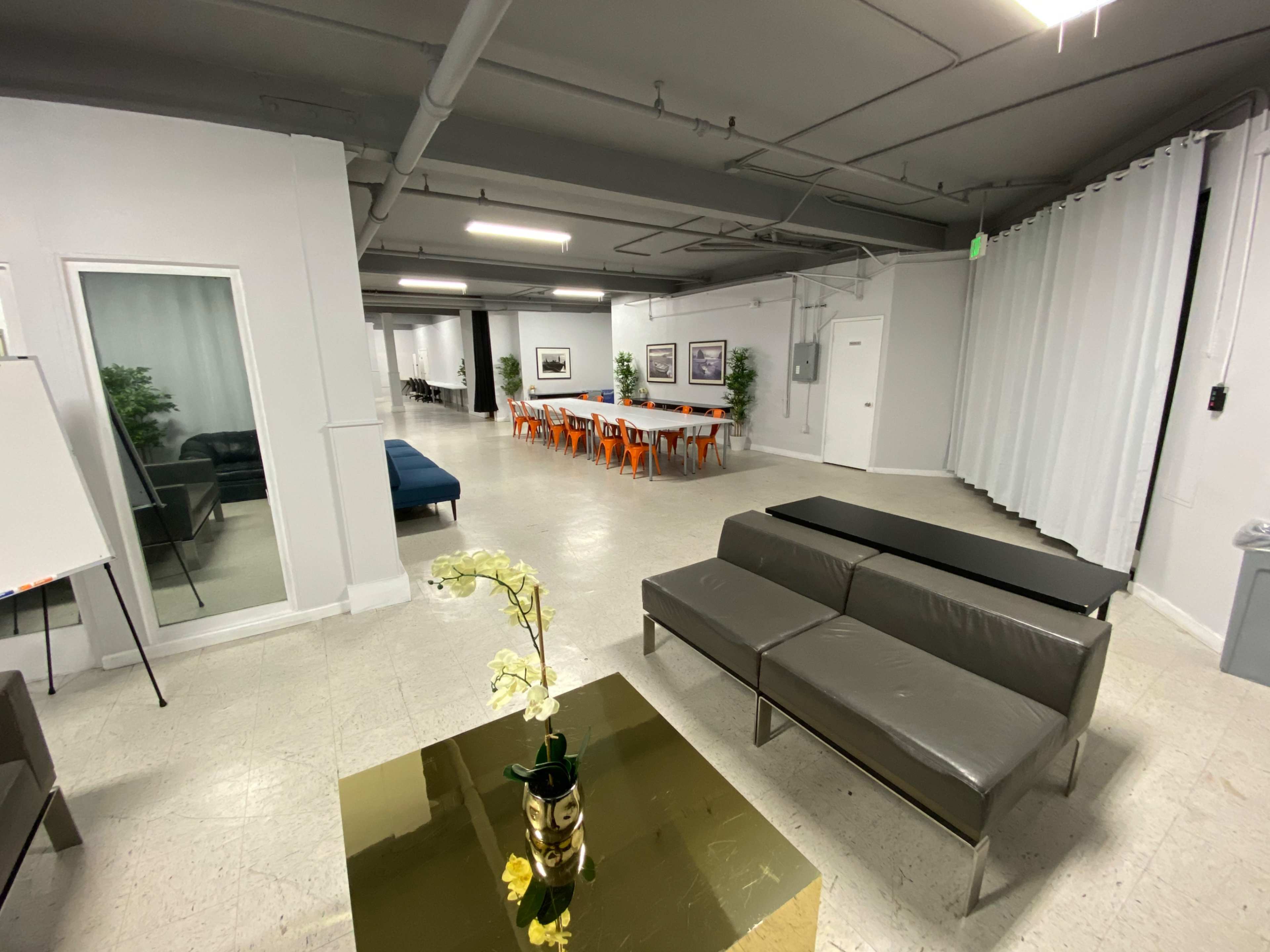 A spacious room with a seating area featuring a gray sofa and glass table, leading to a dining area with orange chairs.