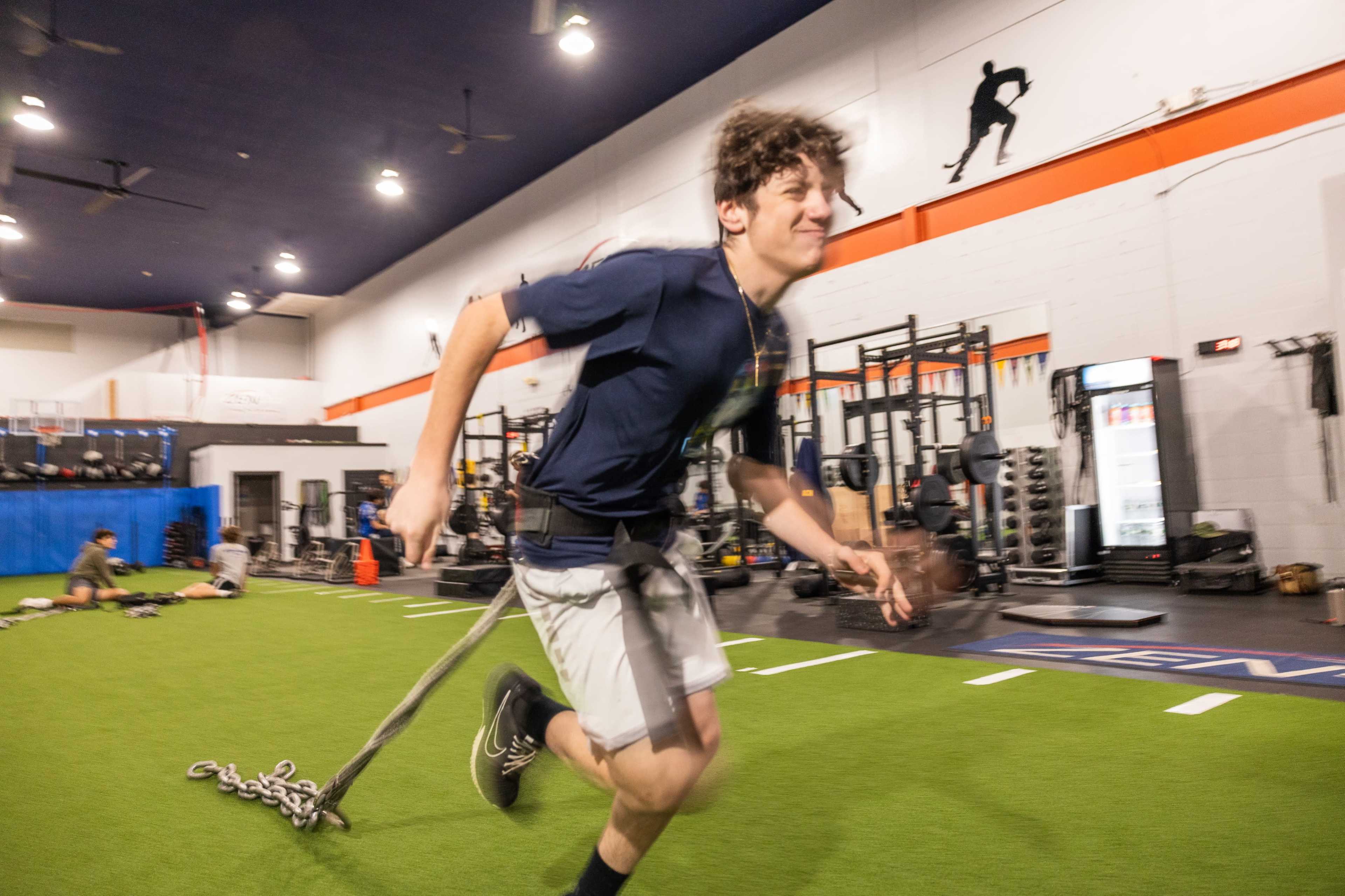 A young man runs on turf while attached to a training harness with chains in a gym equipped with weightlifting stations and fitness equipment.