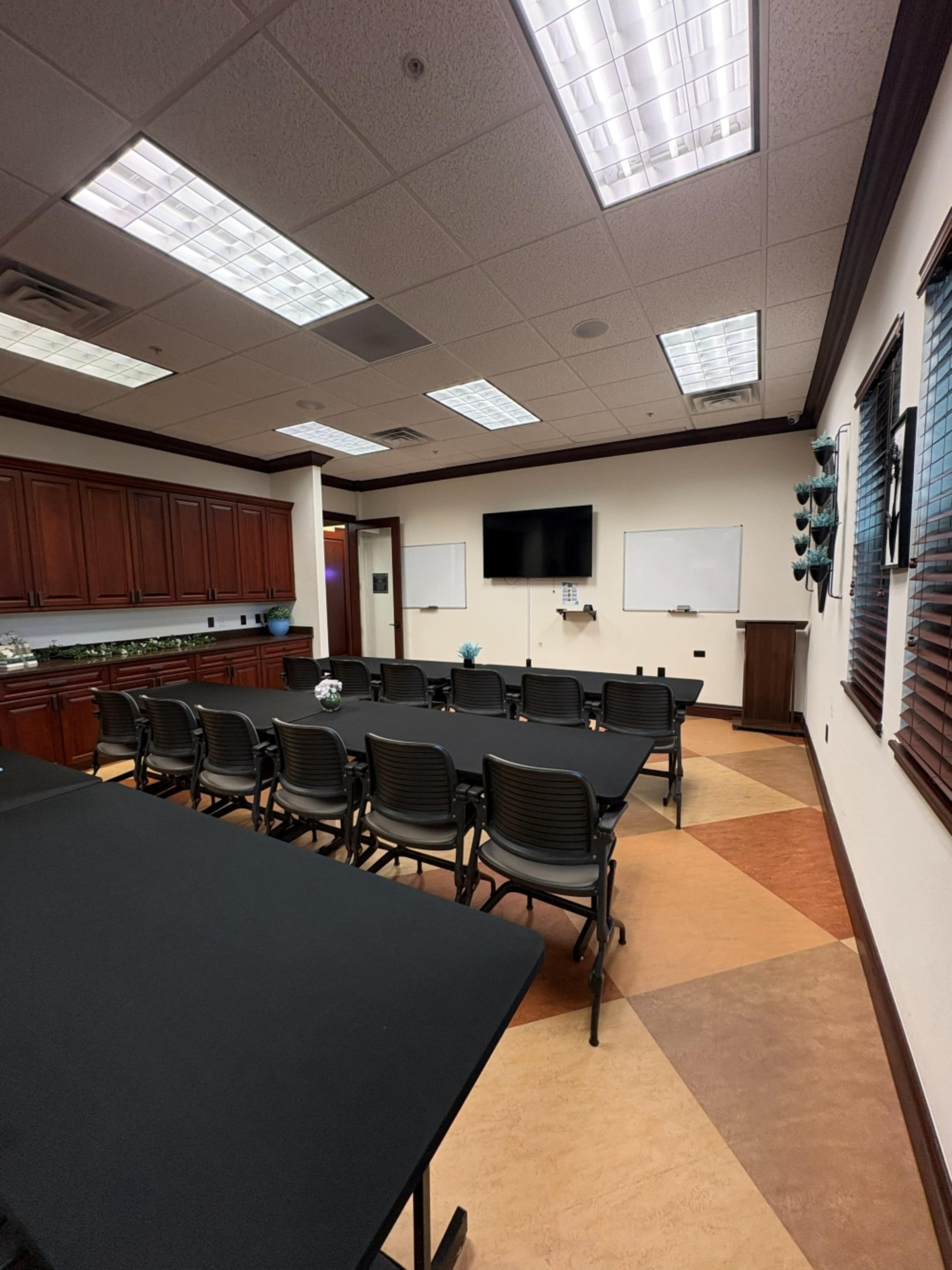 The image shows a conference room with black tables and chairs arranged in a row, featuring a television on the wall and whiteboards on either side.
