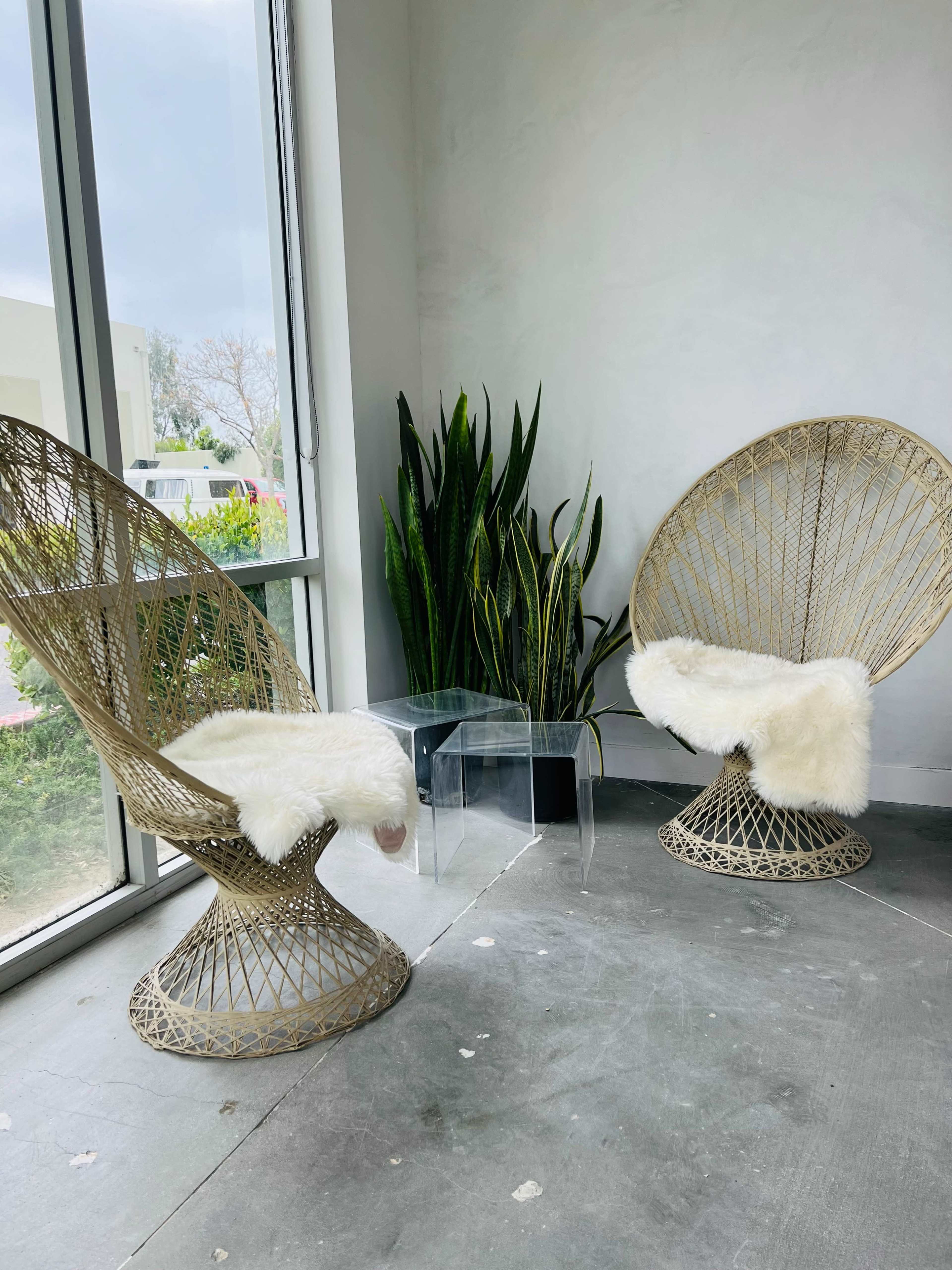 Two woven chairs with faux fur cushions face each other beside a small clear table in a bright, minimalist indoor space.