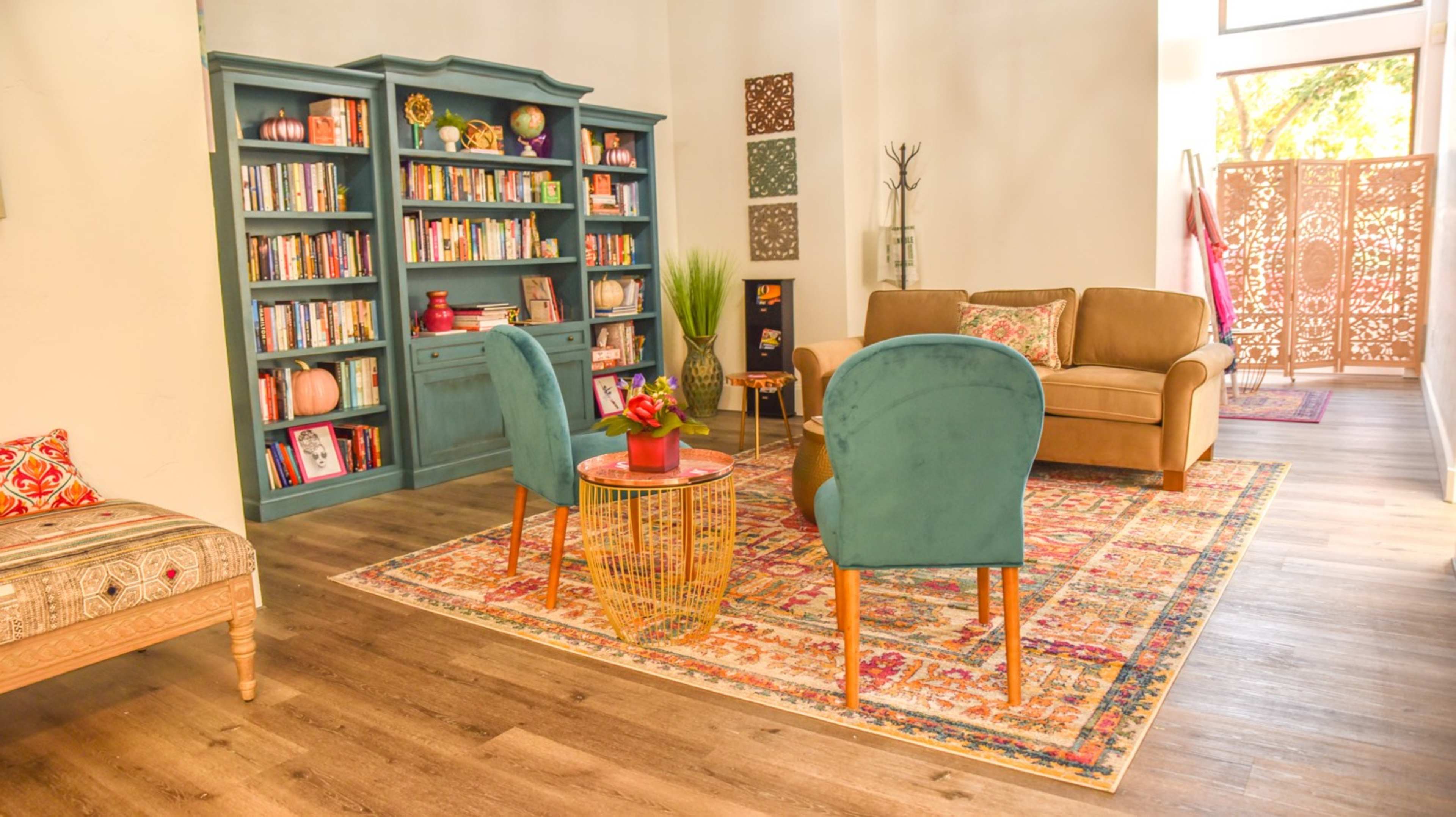 A cozy living room with a blue bookshelf filled with books, a brown sofa, two teal chairs, and a decorative rug on the wooden floor.