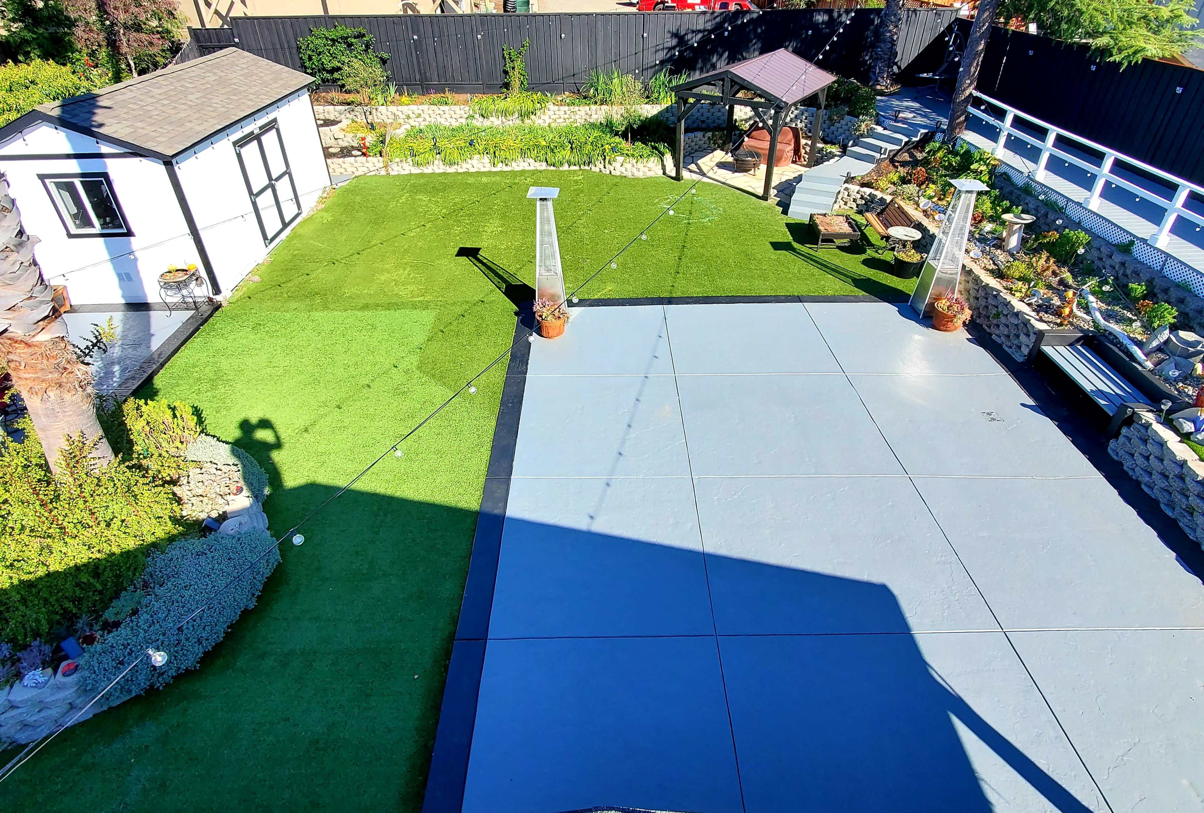 A landscaped outdoor area featuring a gray paving surface, artificial grass, a gazebo, and various garden features including potted plants and a small shed.