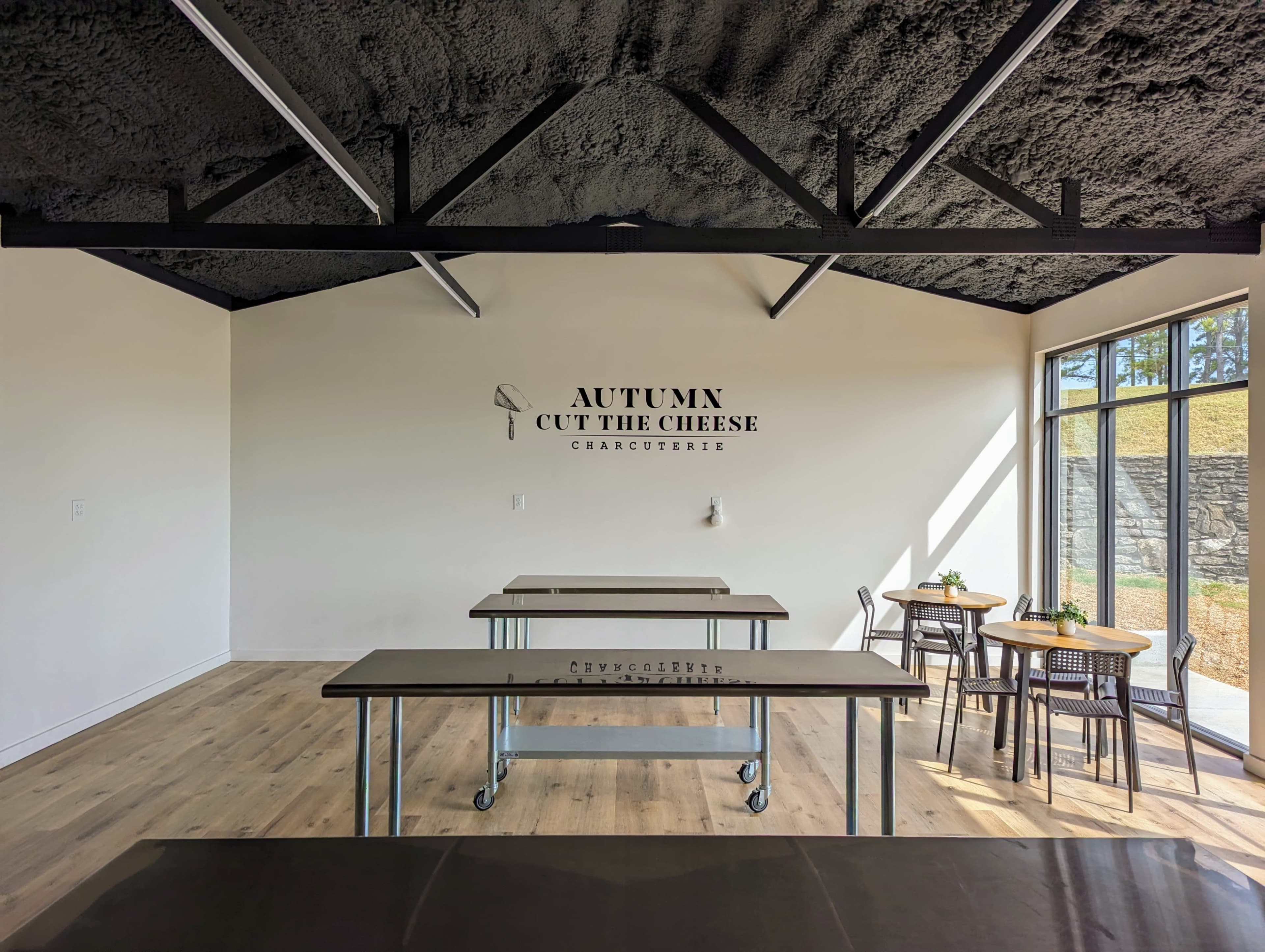 A bright, minimalist space with wooden tables and chairs, featuring a wall sign that reads "AUTUMN CUT THE CHEESE CHARCUTERIE."