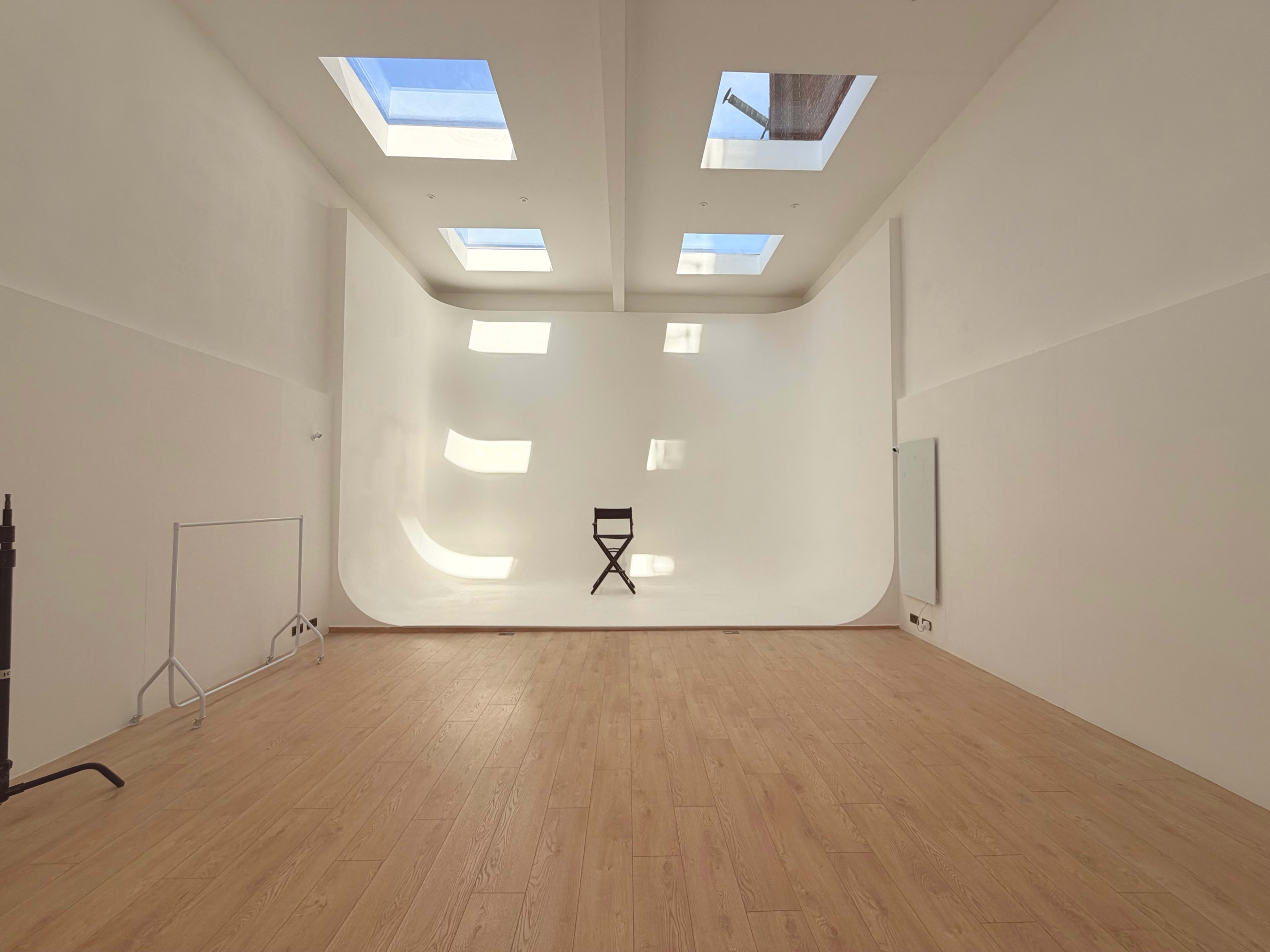 The image shows a minimalist room with a curved white wall, a black director's chair in the center, and several skylights illuminating the space.