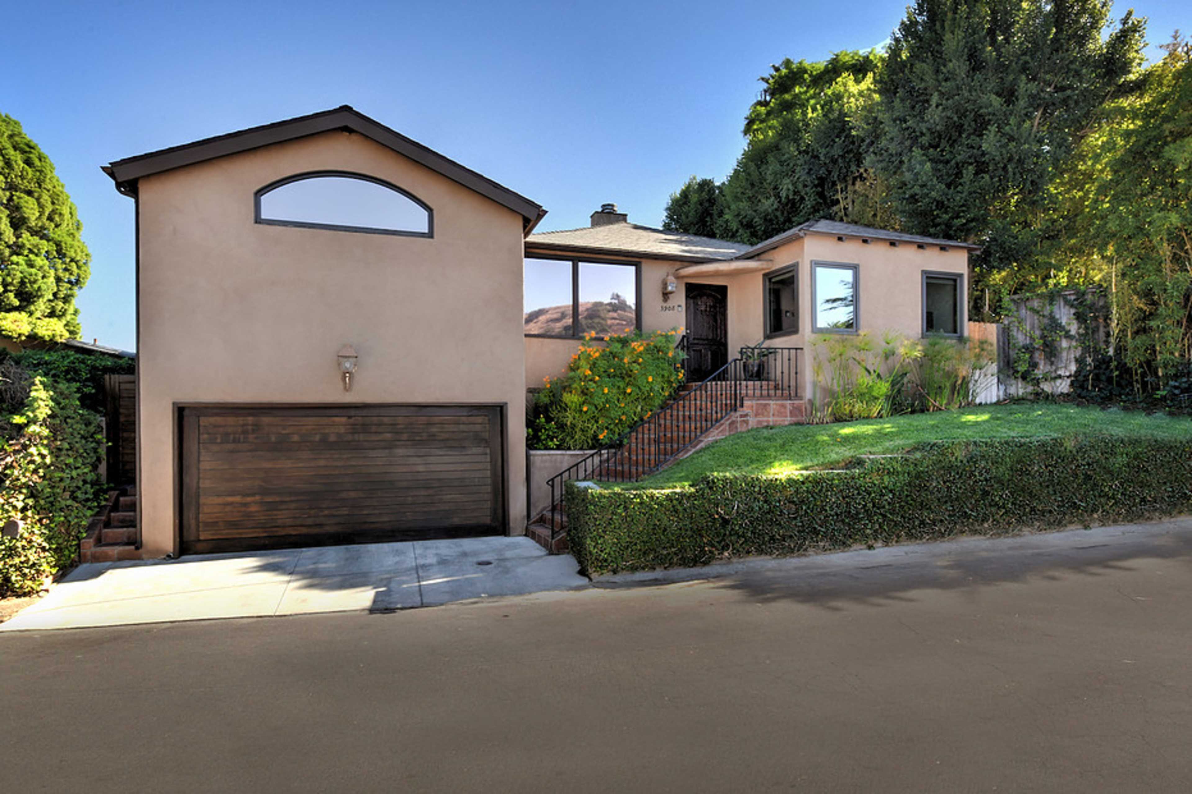 The image shows a two-story house with a garage, surrounded by greenery and set on a sloped street.