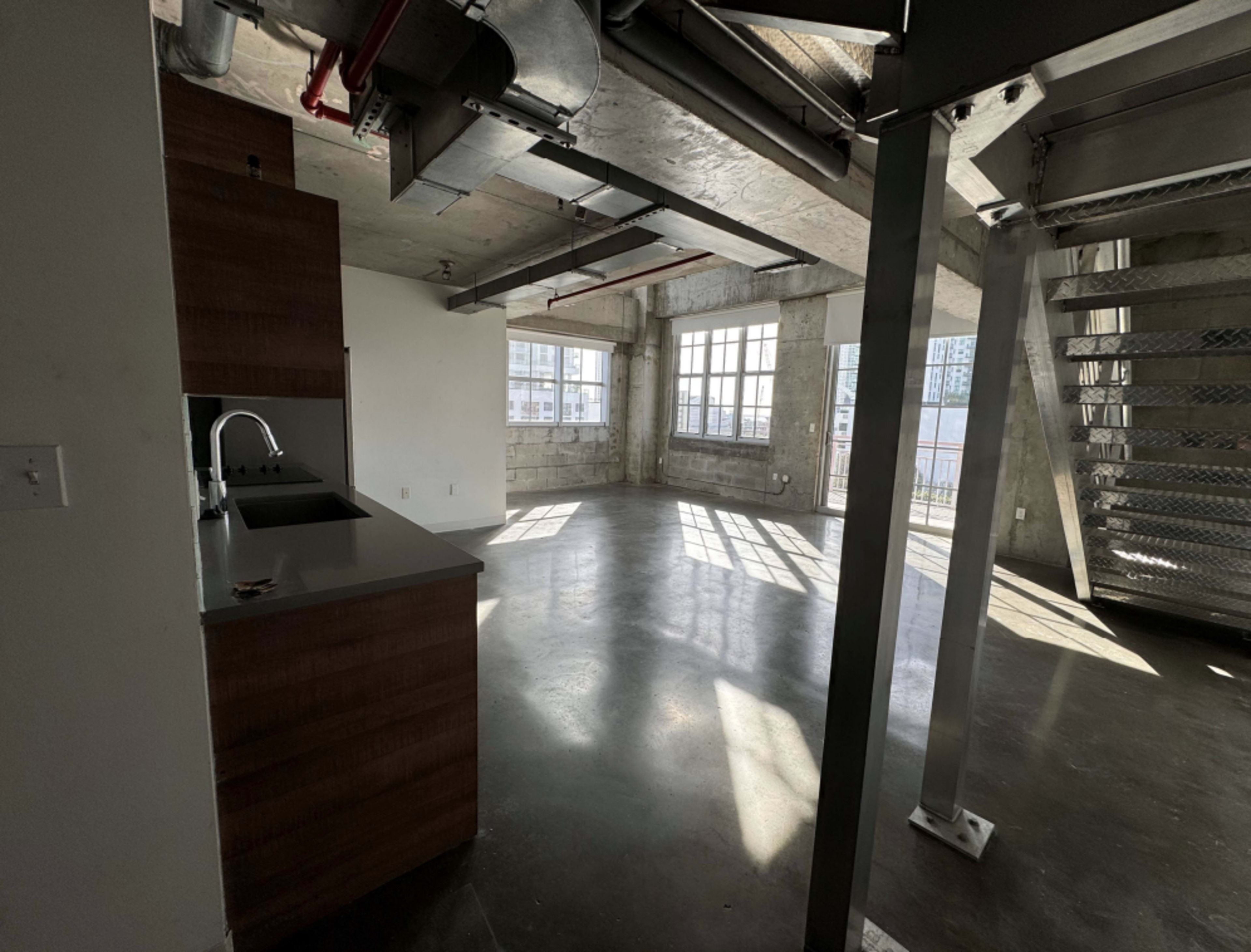 The image shows an industrial-style interior of a living space with concrete flooring and large windows allowing natural light.