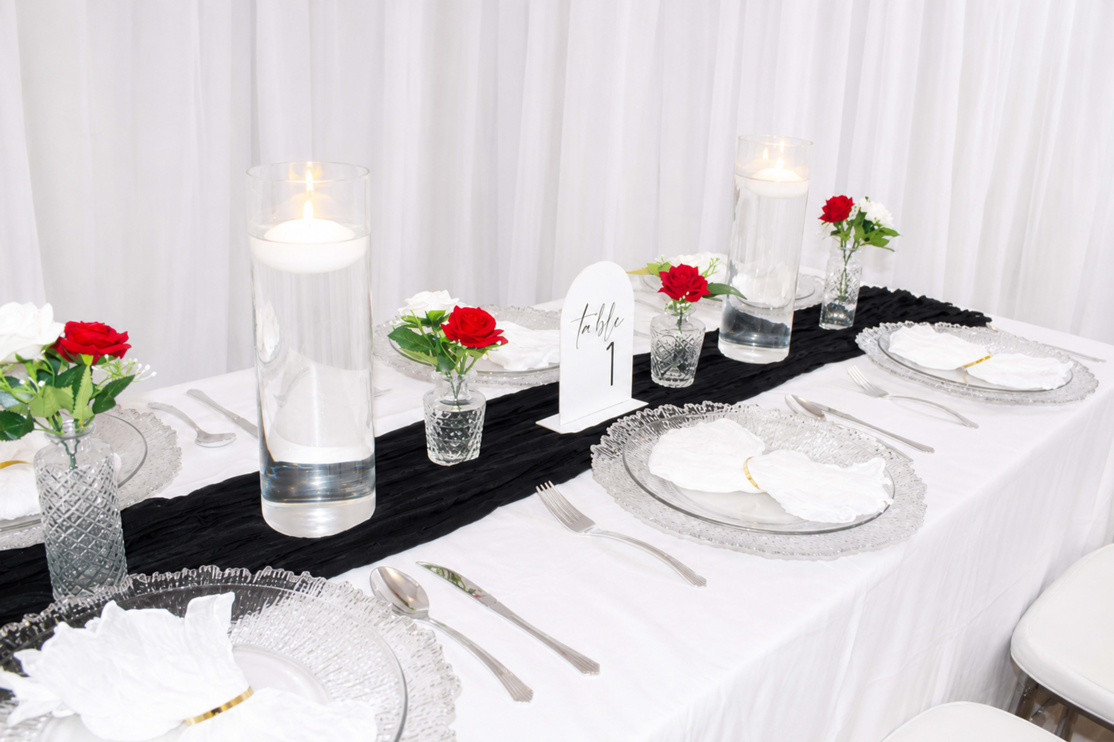 The image shows a elegantly arranged dining table with white tablecloth, crystal plates, glass vases filled with water and candles, and small arrangements of red and white flowers.